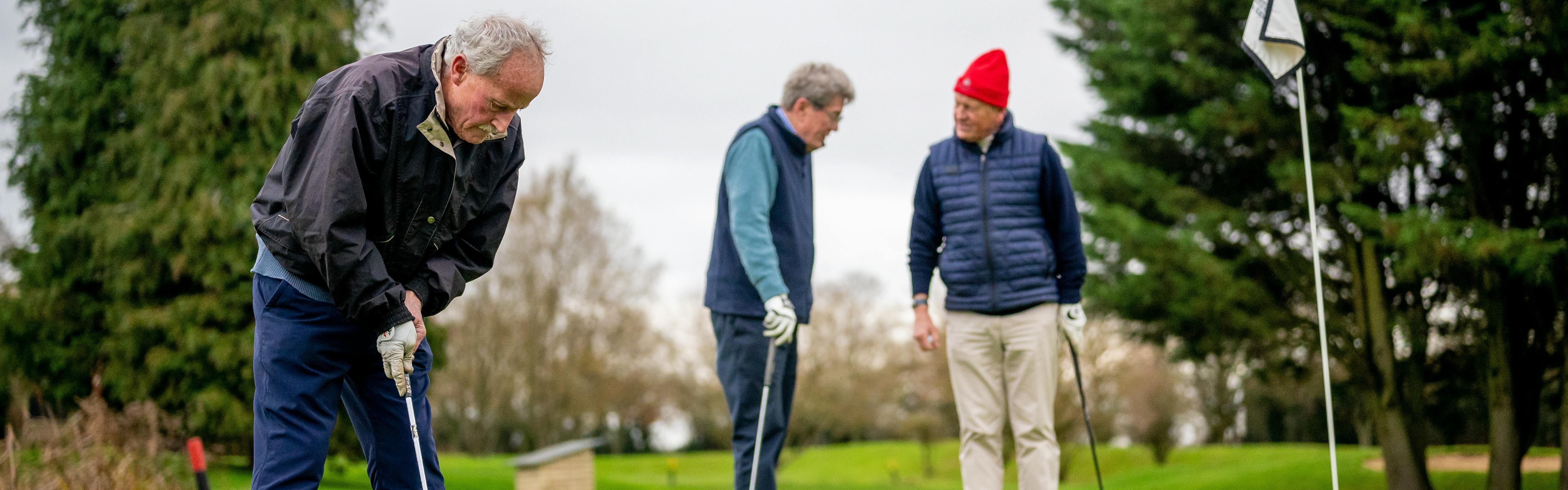 One man lines up to put the ball into the hole as two men stand and talk in the background. 