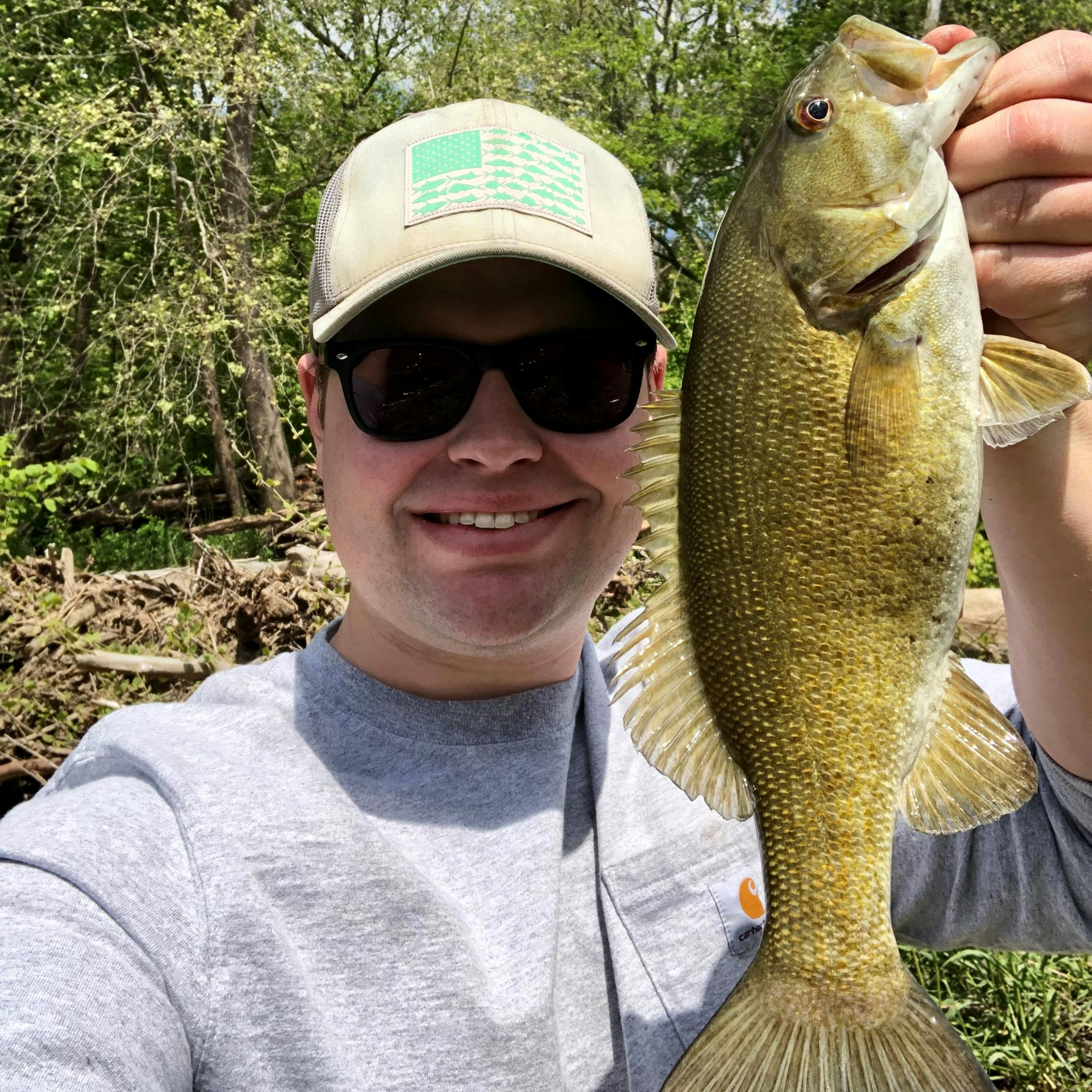 A man with a hat and glasses holding up a smallmouth bass. The fish is green.