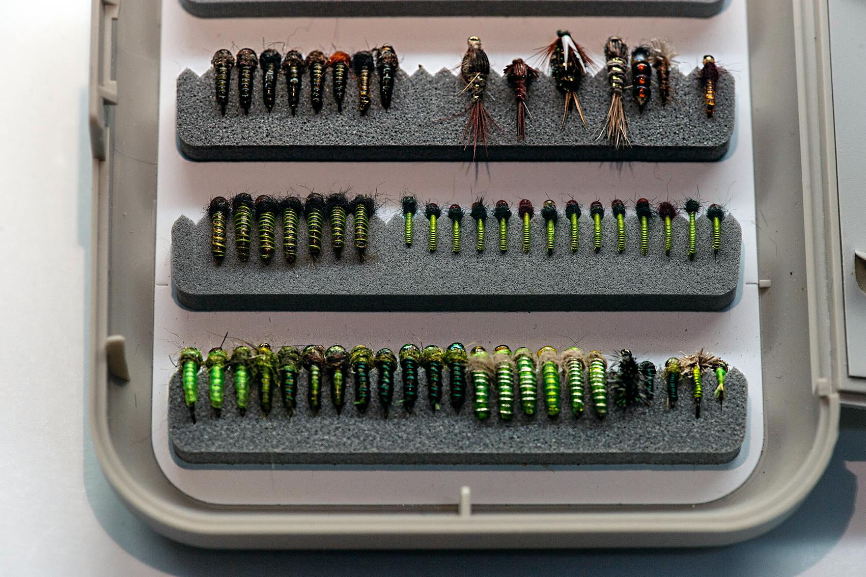 Several different types of flies for fly fishing sit in a fly box. There are three rows with some brown flies, some black flies, and some green flies.