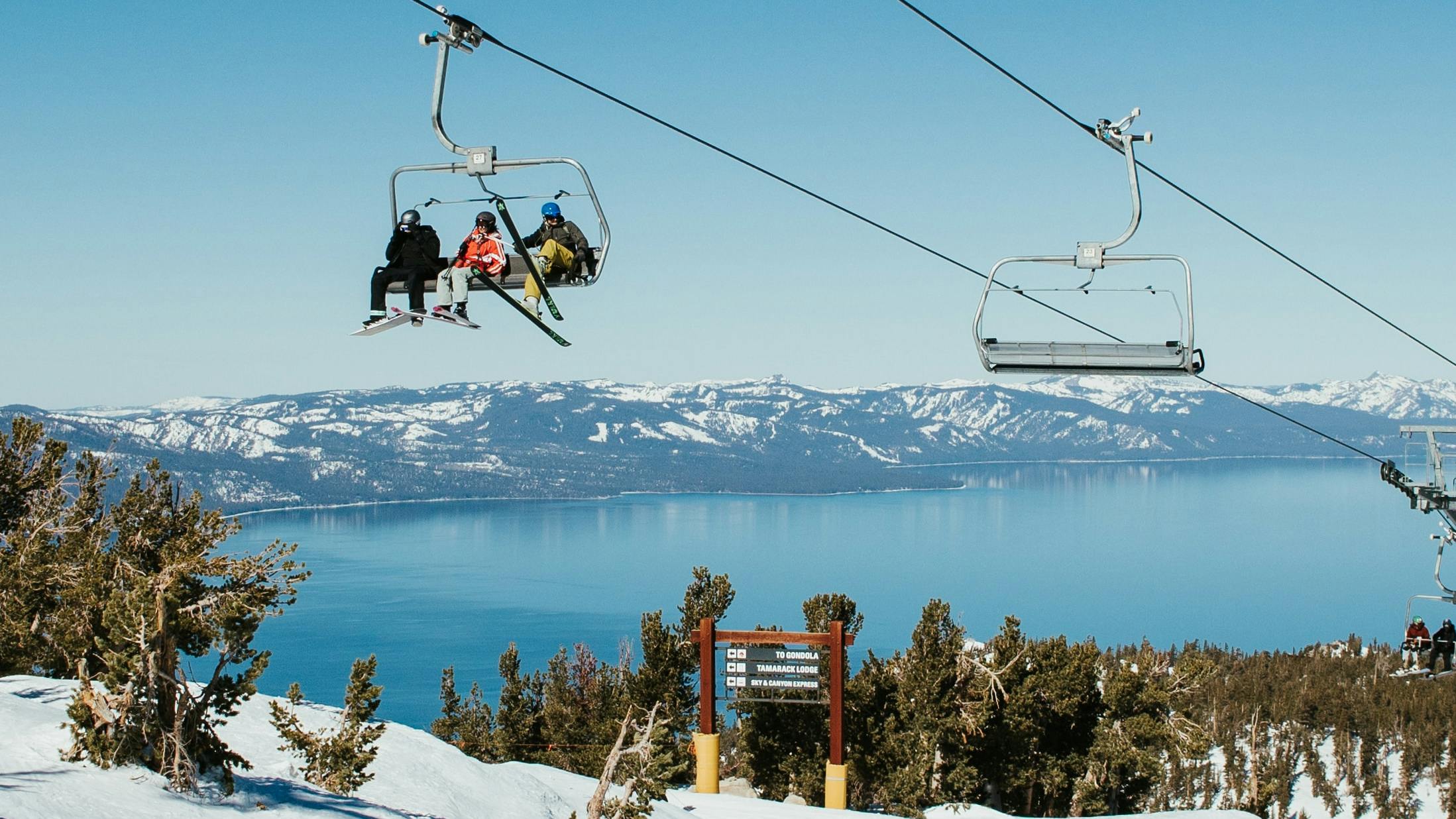 Skiers sitting on a chairlift with Lake Tahoe in the background. There are snowy mountians surrounding the lake. 