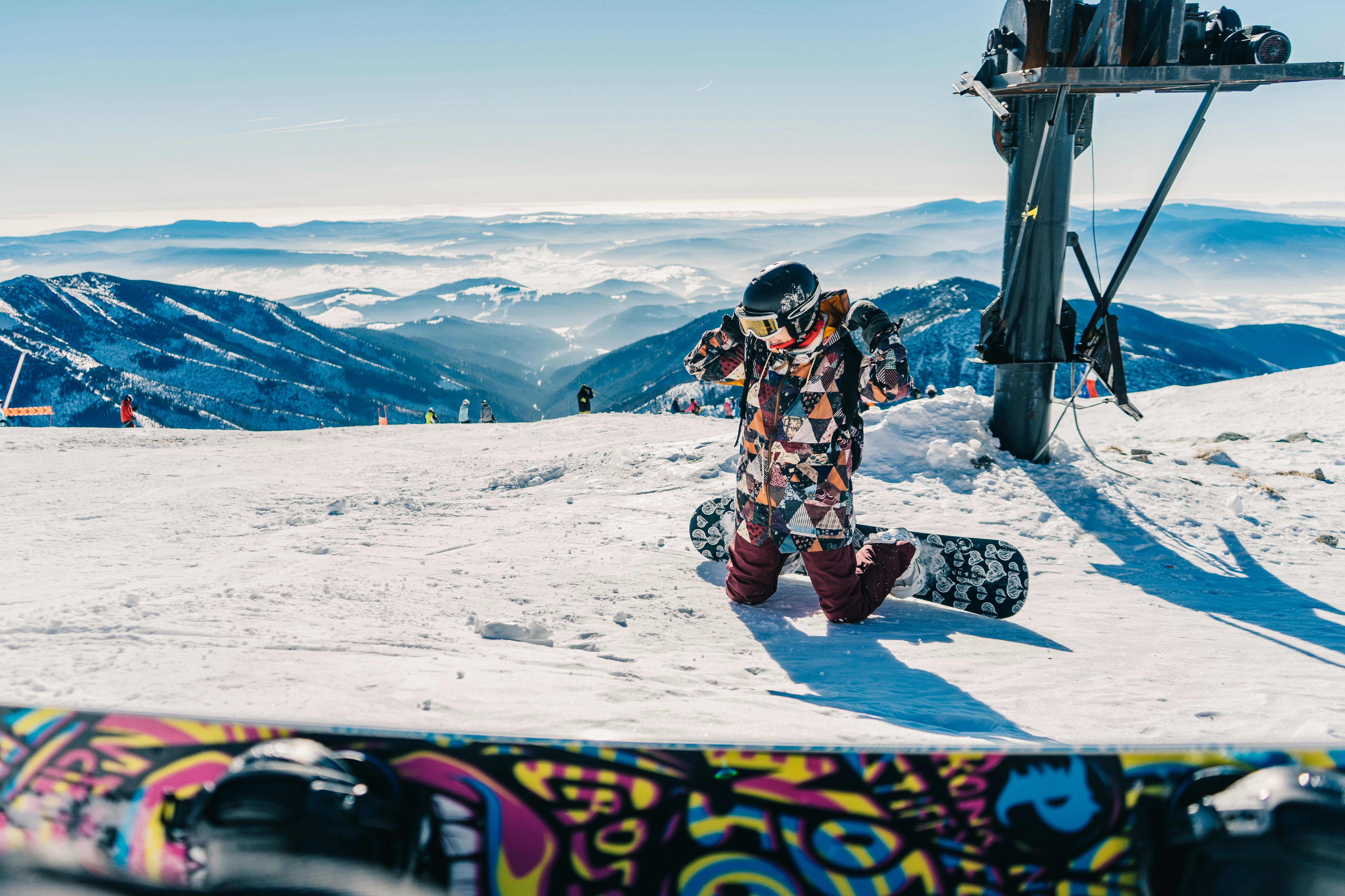 A snowboarder in a brightly colored geometric pattern jacket kneels to adjust their hood