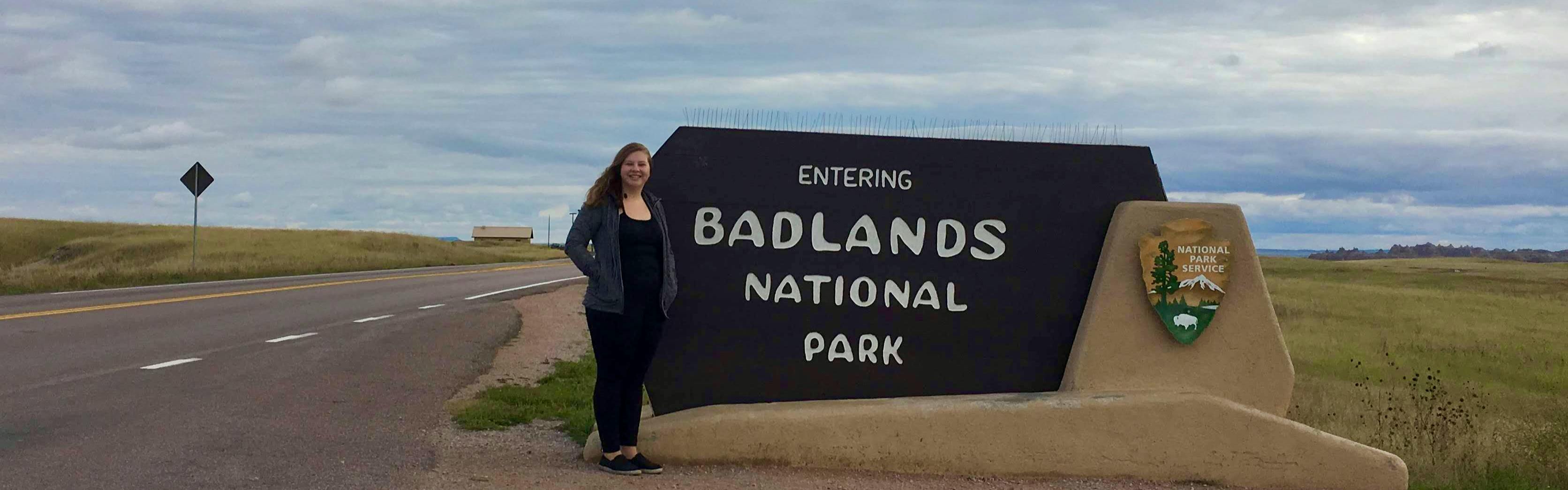 The author stands on the side of the road next to a National Park Service sign that reads Entering Badlands National Park. 