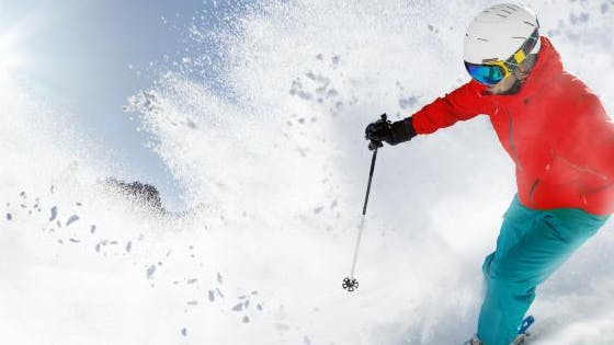 A skier in a red jacket and turquoise pants skiing down a slope through powder snow