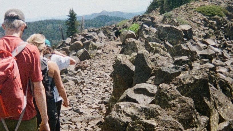 Three hikers walk along a rocky trail. They are all wearing backpacks. 