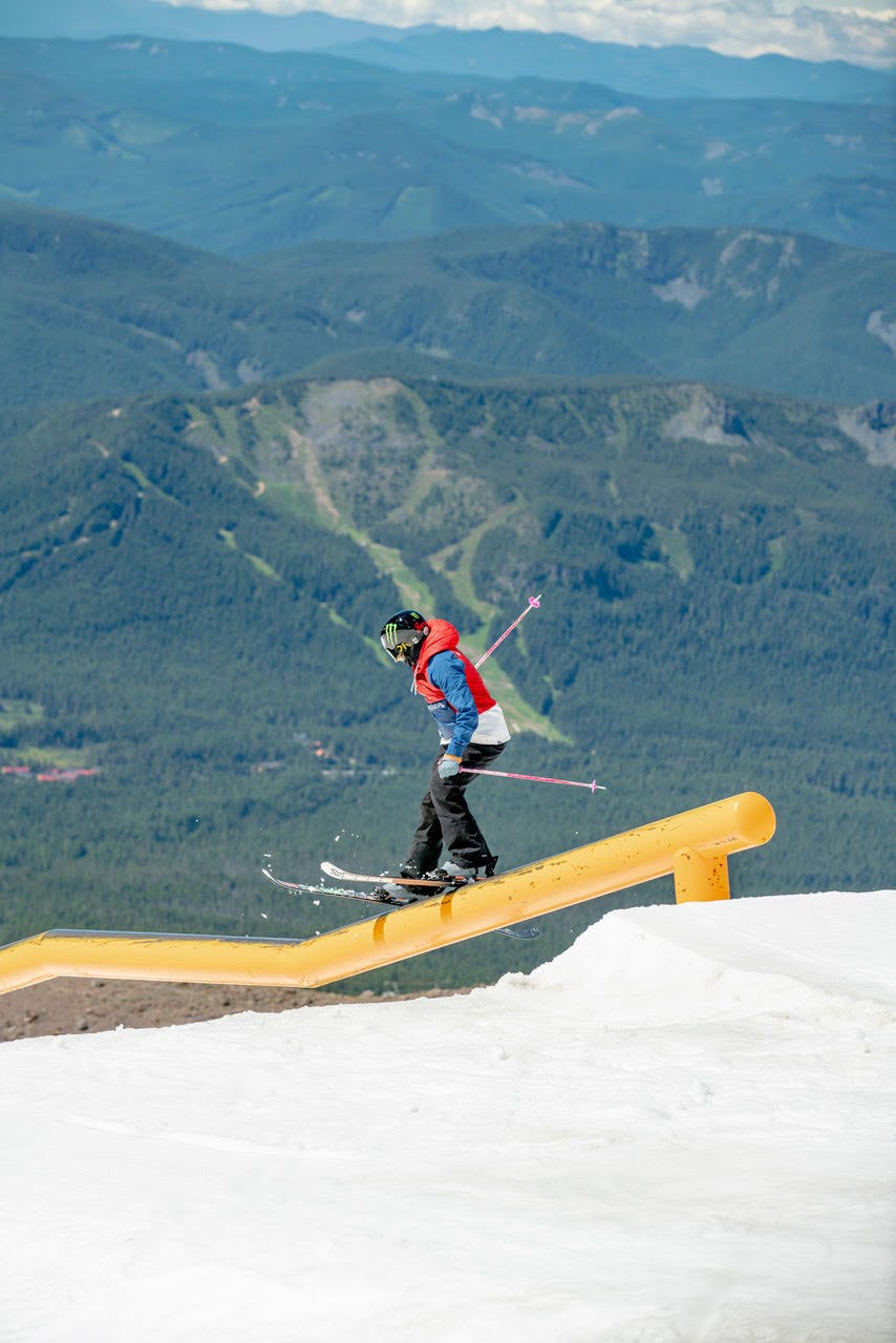 Maggie skis down a yellow rail. Green, tree-covered hills are visible below her. 