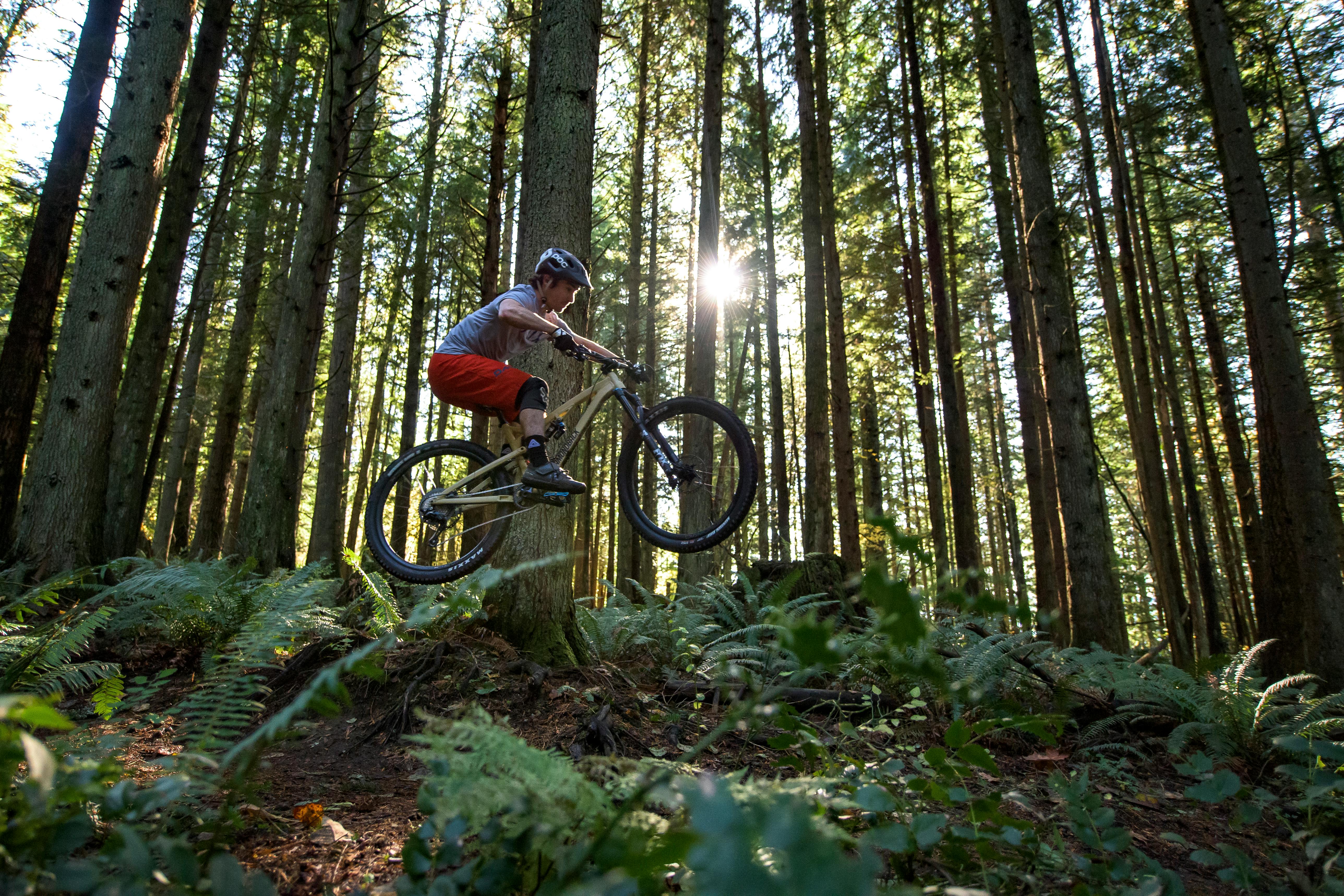 Man airborne on his Diamondback mountain bike after launching off a jump in the forest. The sun shines through the trees.
