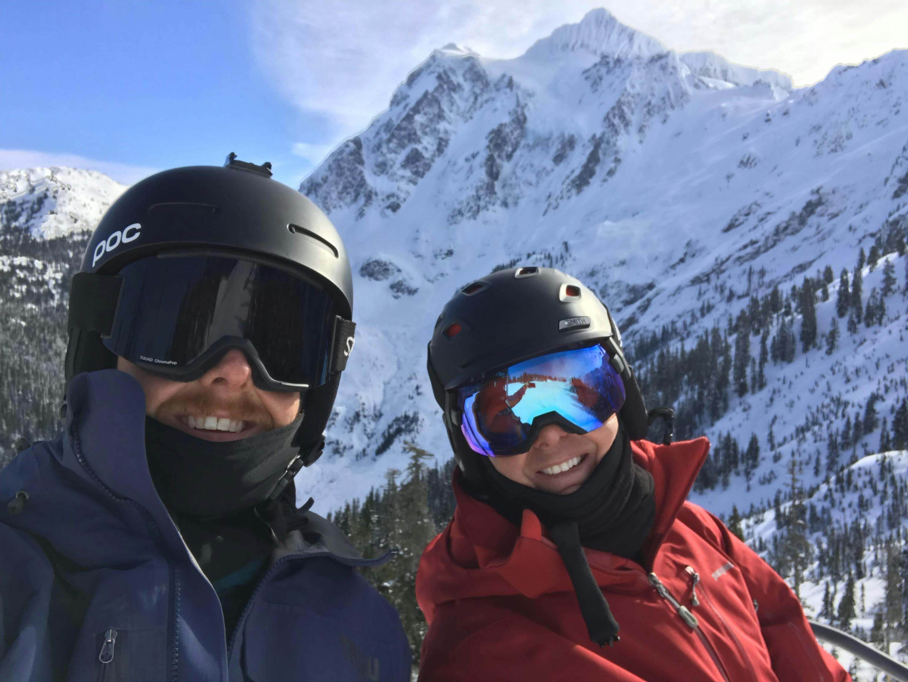 Two skiers, one with a very dark lens and the Smith Squad goggles. There is a snowy mountain range in the background.