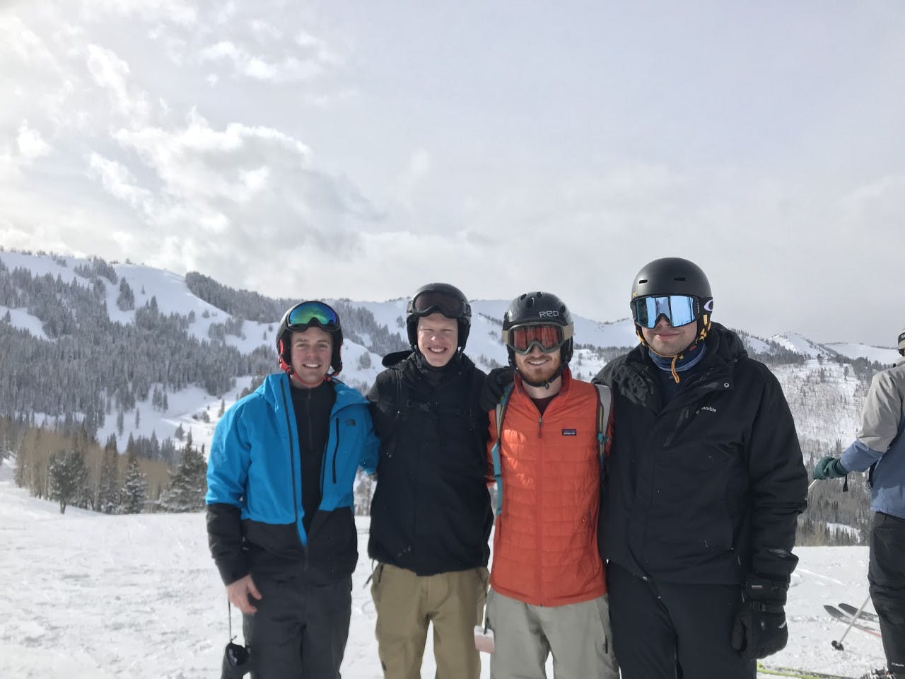 Four people standing at the top of a ski run. They all have their ski gear on and there is a snowy mountain in the background. 