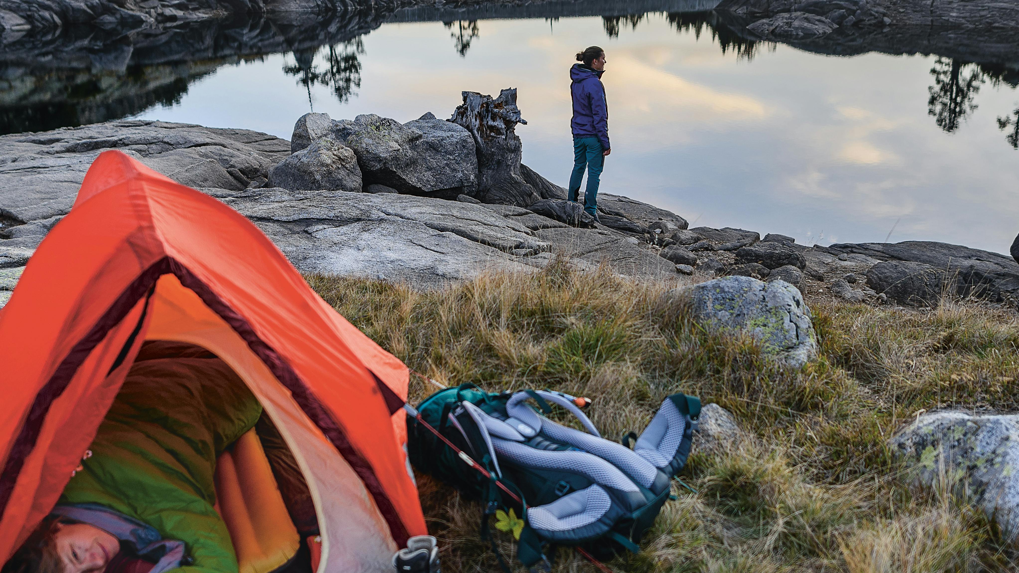 A camper standing by a lake while another camper cozies up in her sleeping bag in an orange one-person tent