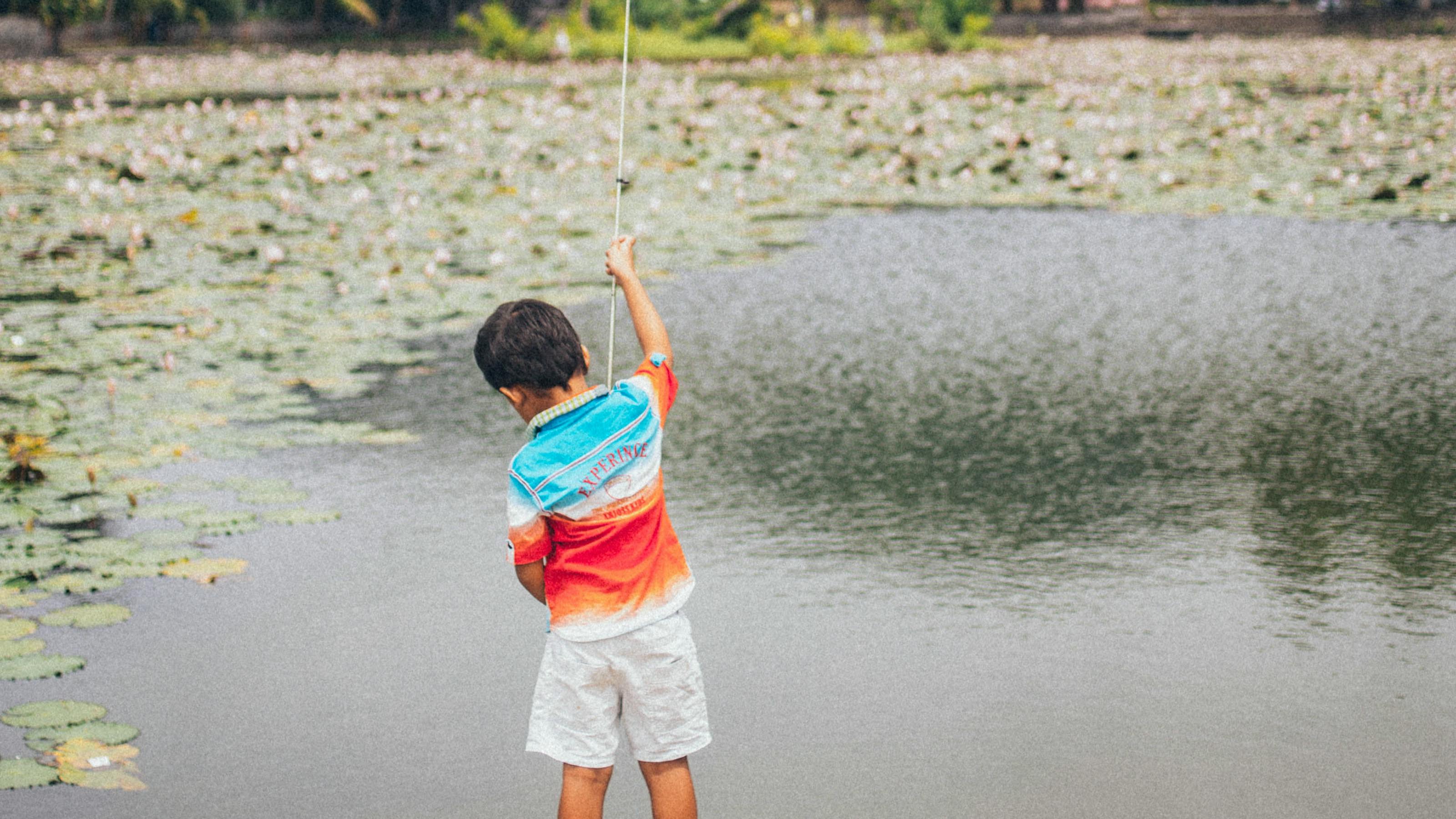 A young boy holds a fishing rod in front of a pond with water lilies on it. 