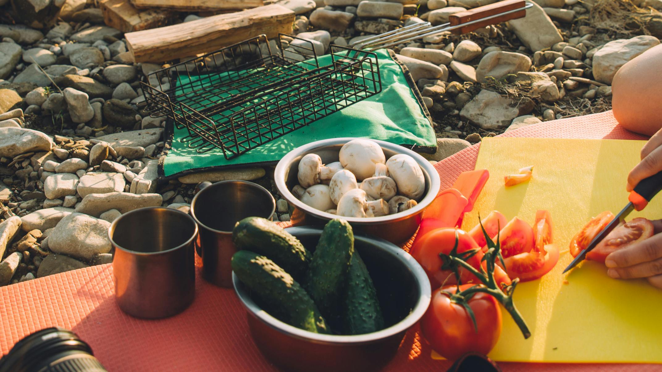 A person cuts up a tomato while sitting on a rocky surface. Two bowls full of pickles and mushrooms also sit there