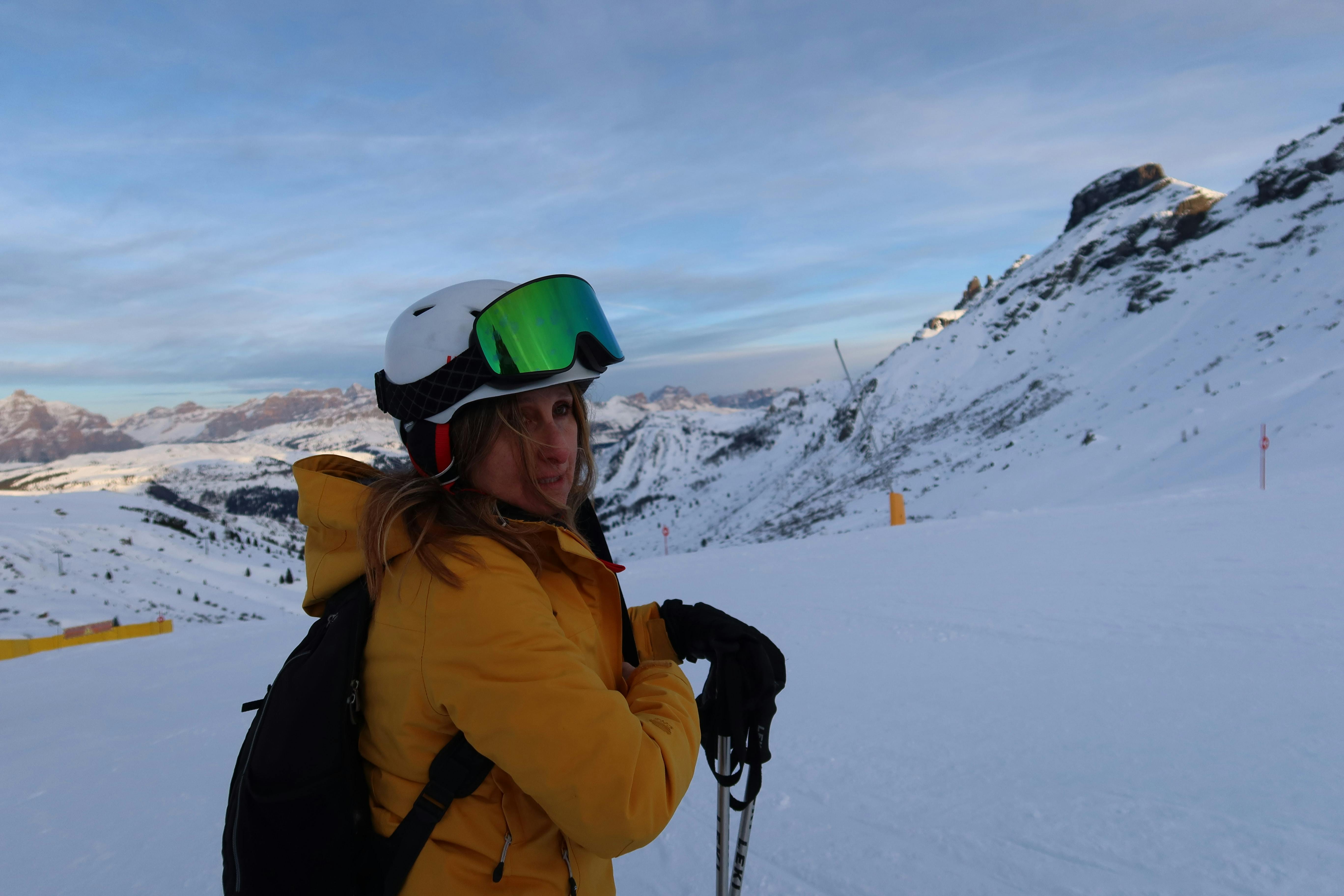 A skier standing in the snow with a helmet and goggles on. She is looking at the camera and there is a snowy mountain in the background. 
