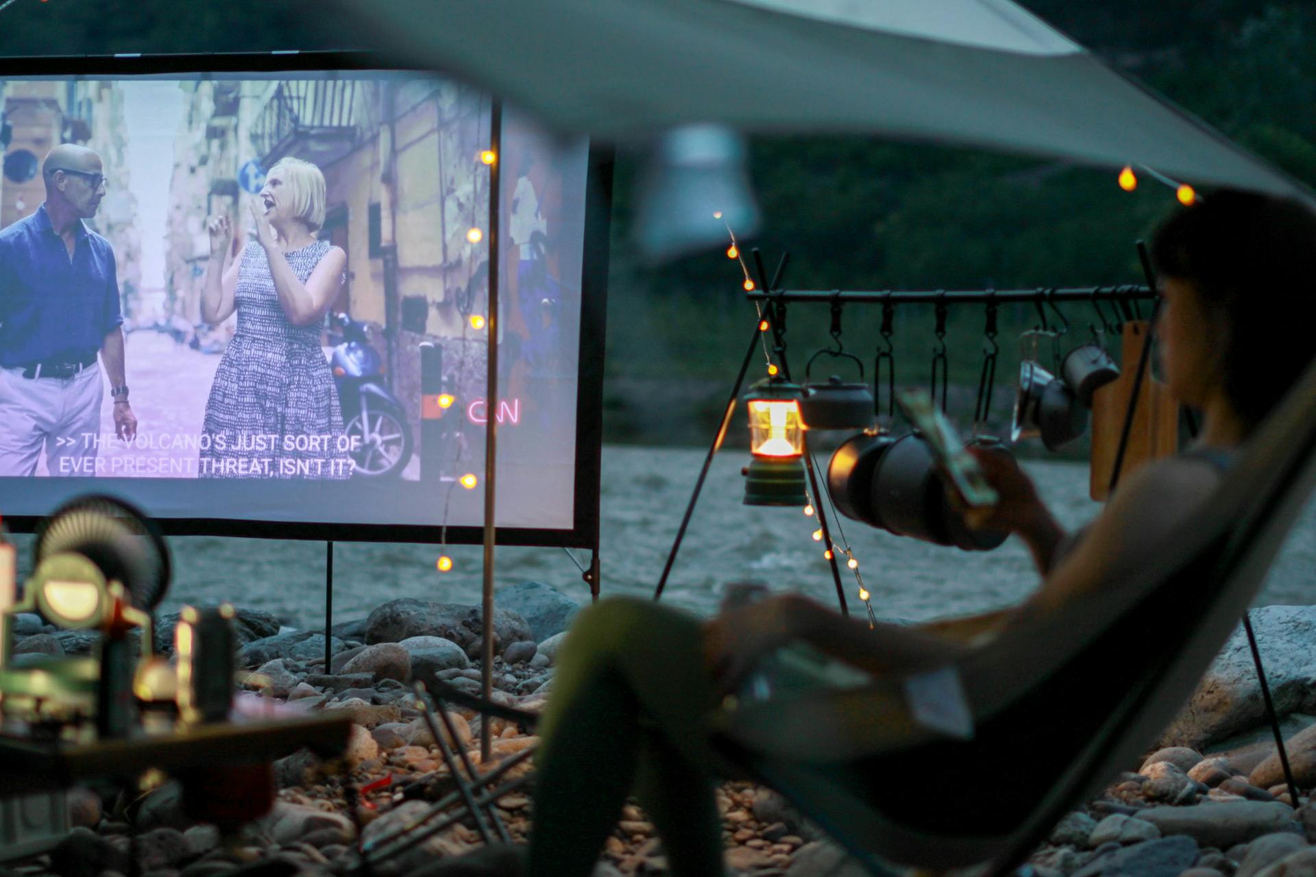 A person sits in a camp chair outside with a projector on a screen. There are lights and blankets around.