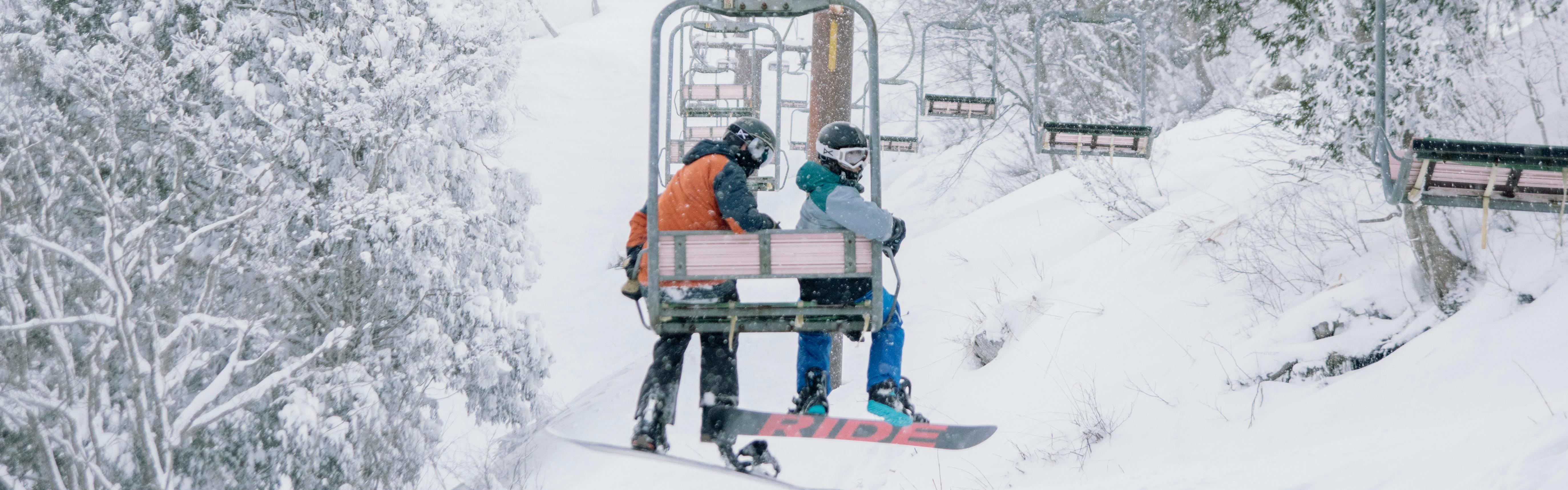 Two people ride a chairlift on snowboards and look back to the camera behind them. 