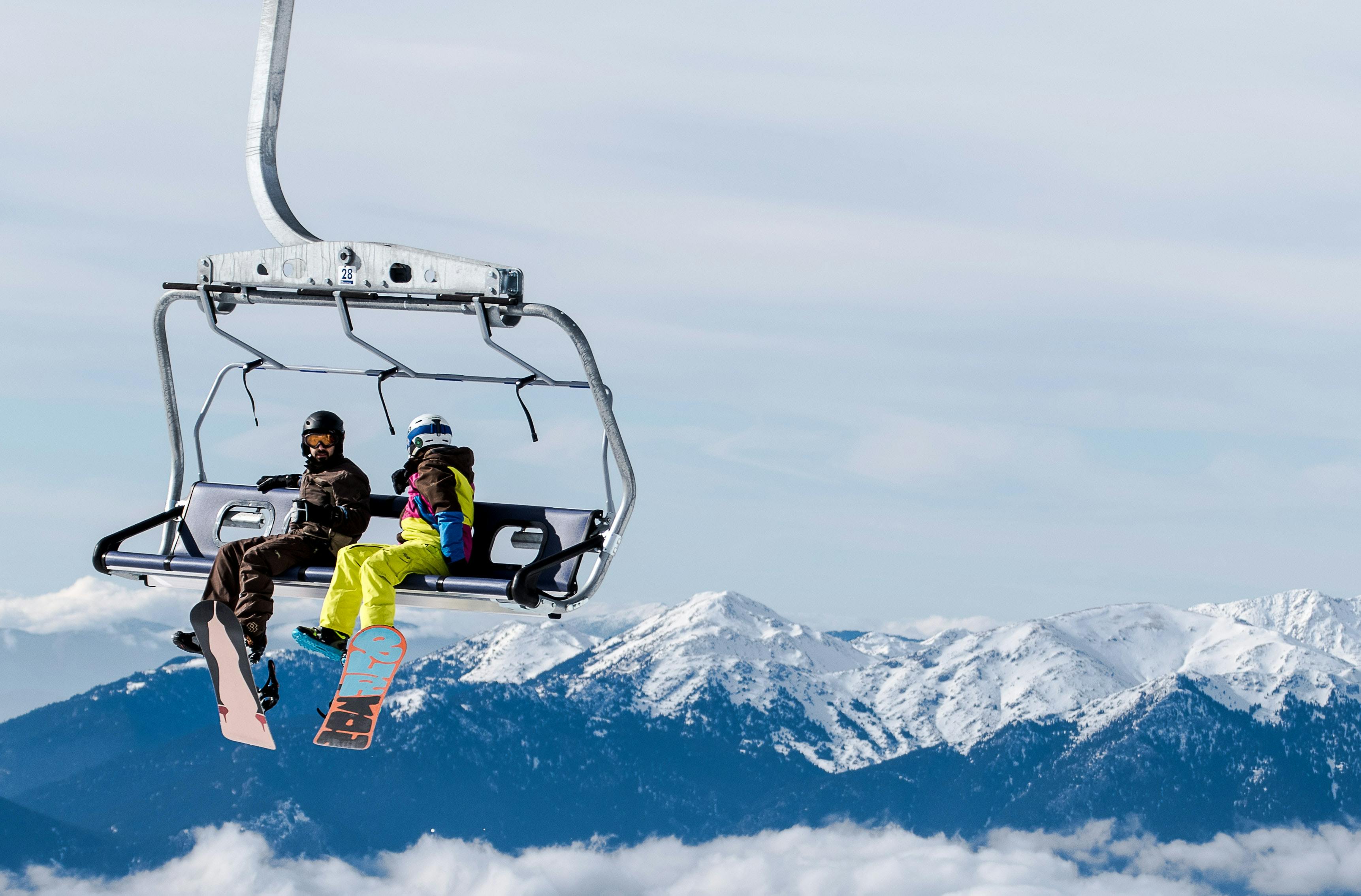 Two snowboarders sit on a chairlift. The ground is not visible below them to they appear to be floating. A snowcapped mountain range extends across the background. 