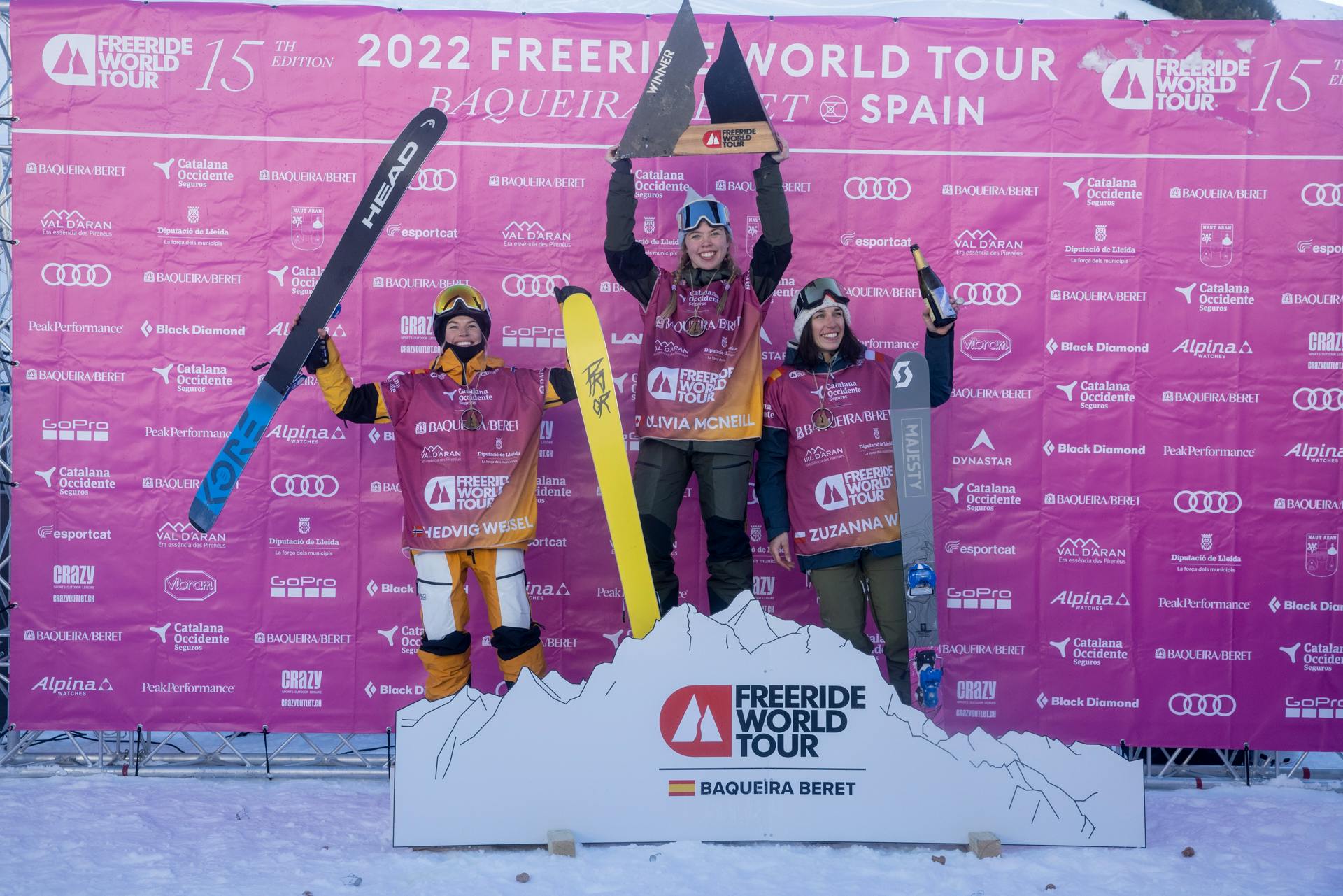 The top three female skiers stand on a podium and hold up their skis and trophies against a pink FWT background. 