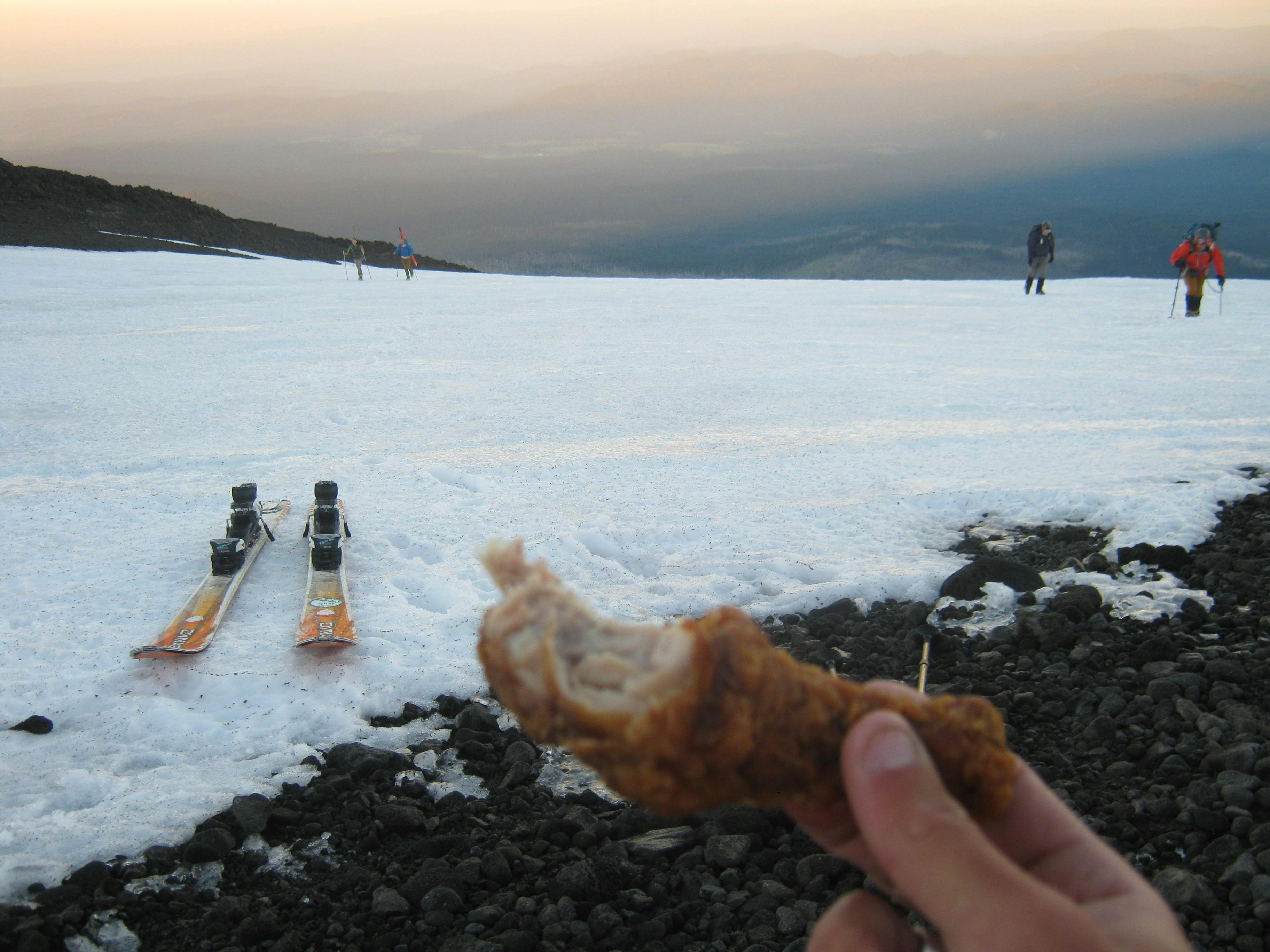 A pair of skis laying on a snowy area. There is a sunset in the background and a hand holding a piece of fried chicken in the corner of the photo. 