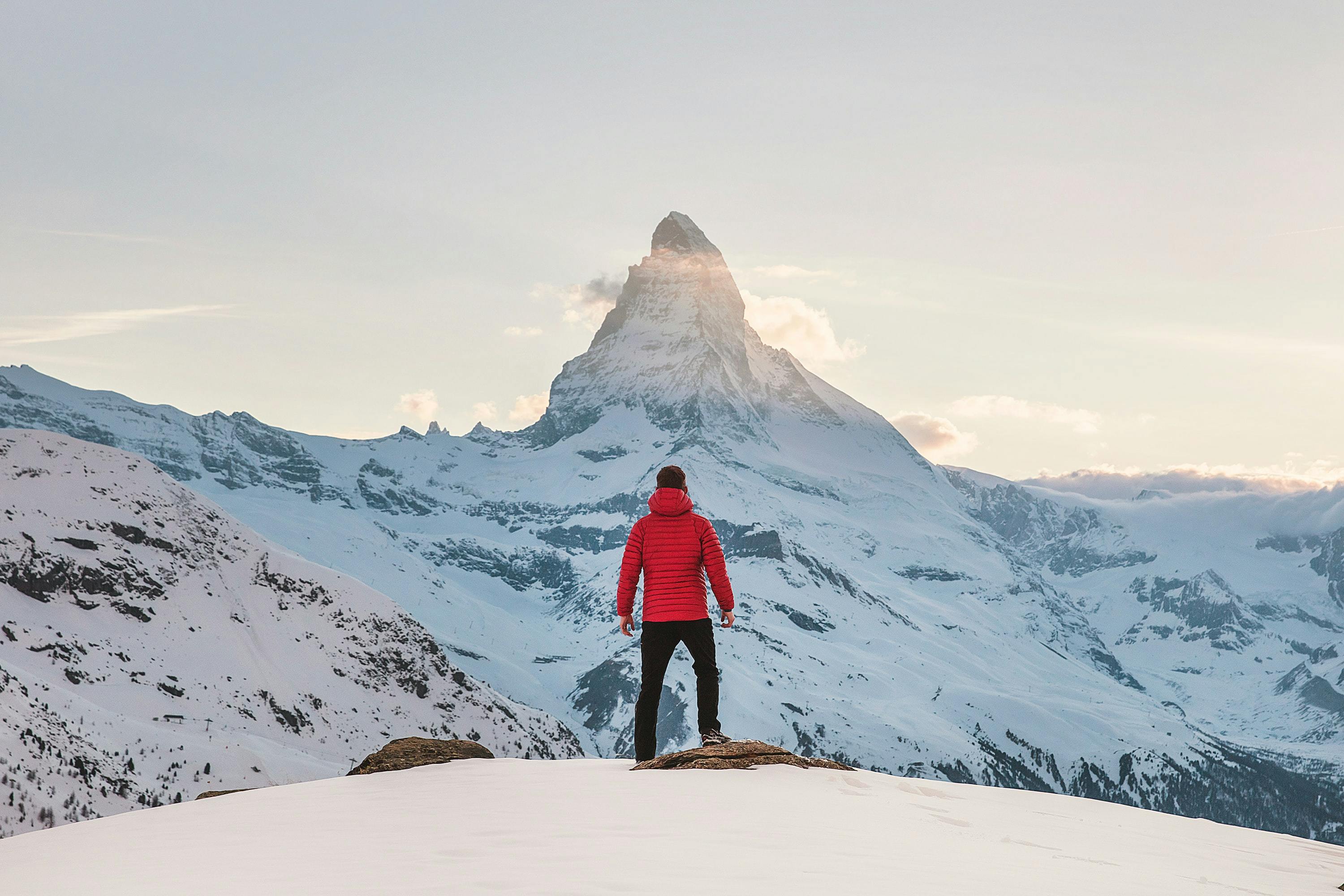 A man in a red jacket and black pants looking out at the snowy mountains before him