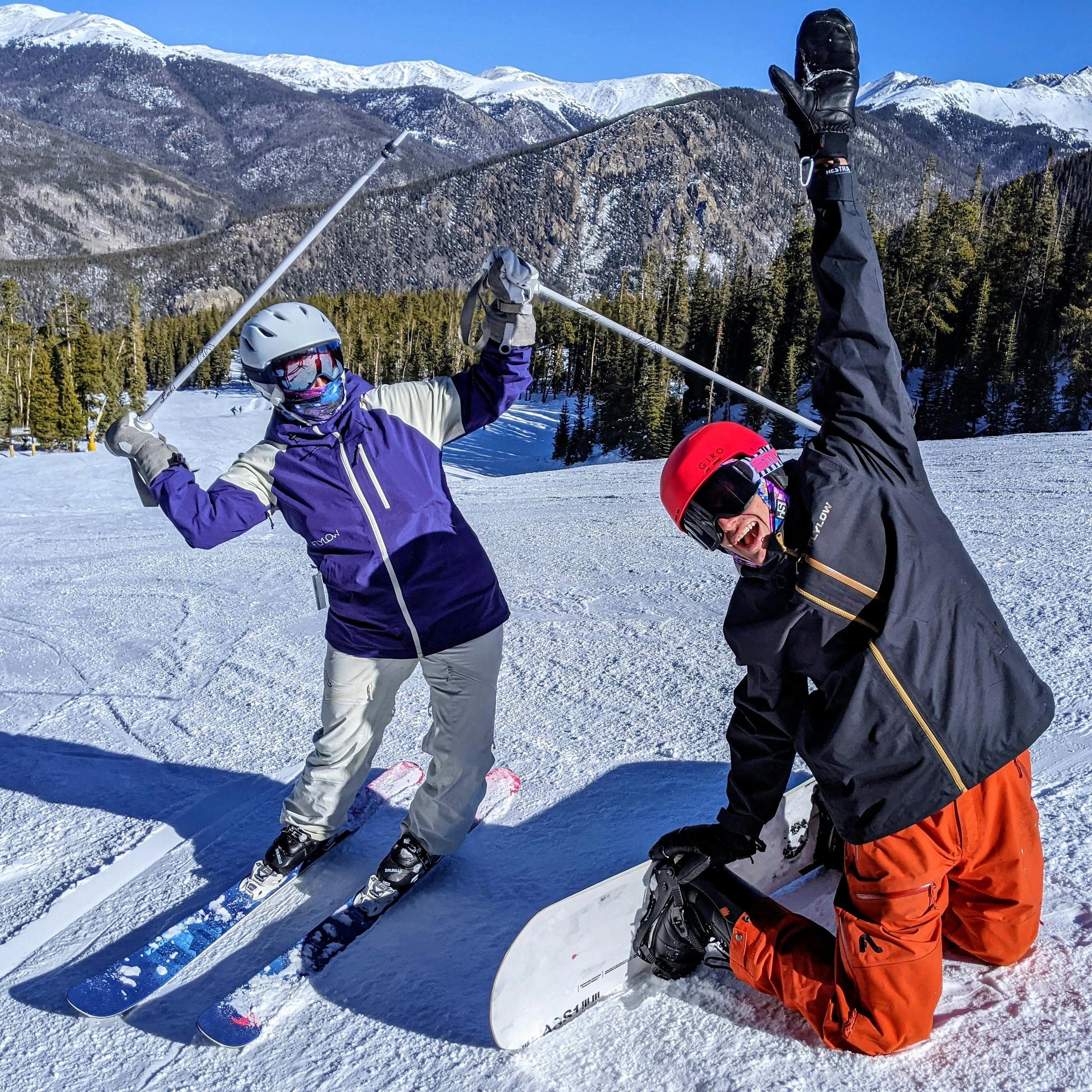 Two people on a ski run, one skier and one snowboarder. They are both looking at the camera with their hands up. 