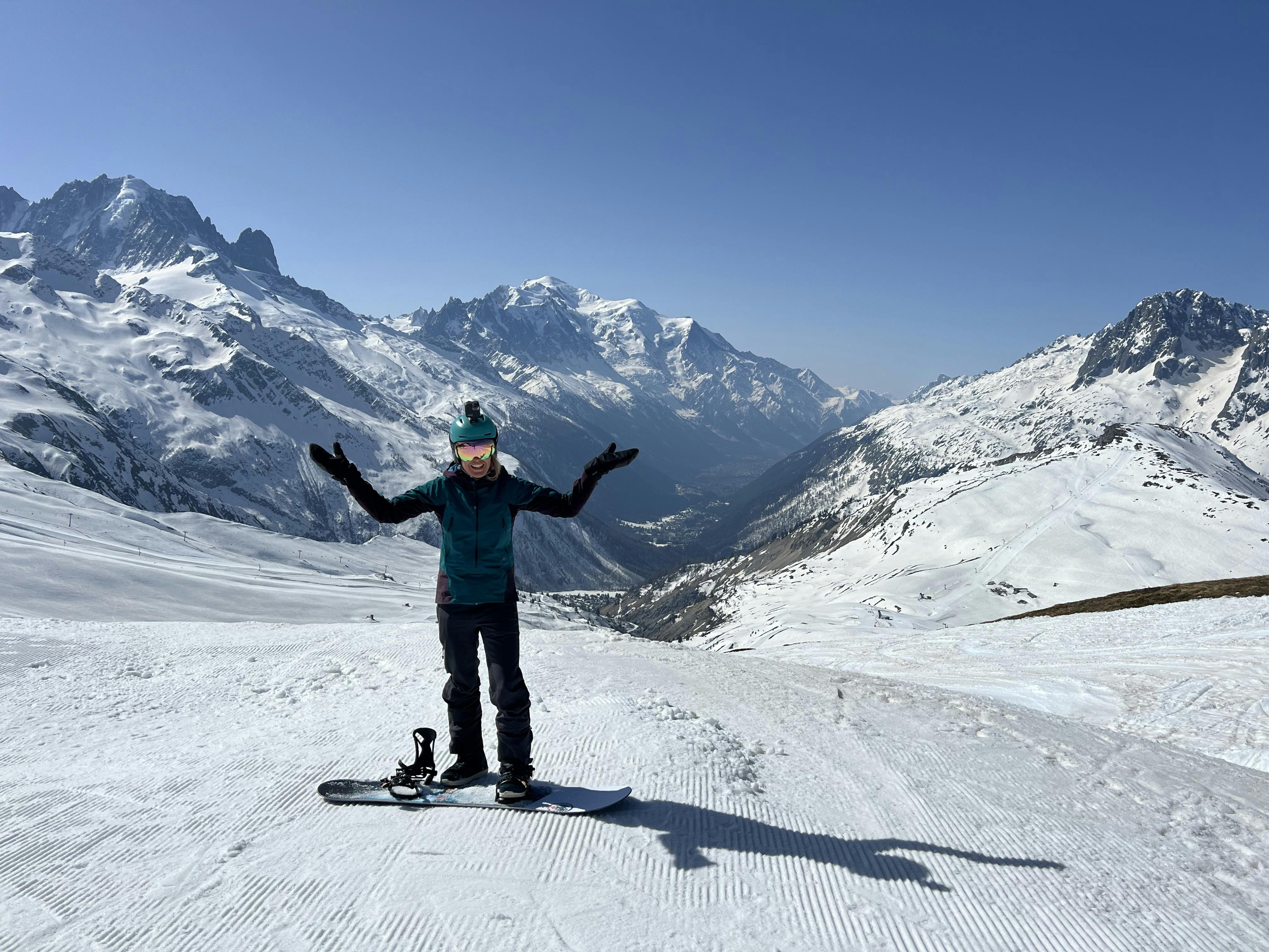 A woman stands on a ski resort trail holding both her hands up. Her snowboard is next to her and there is a mountain range behind her.