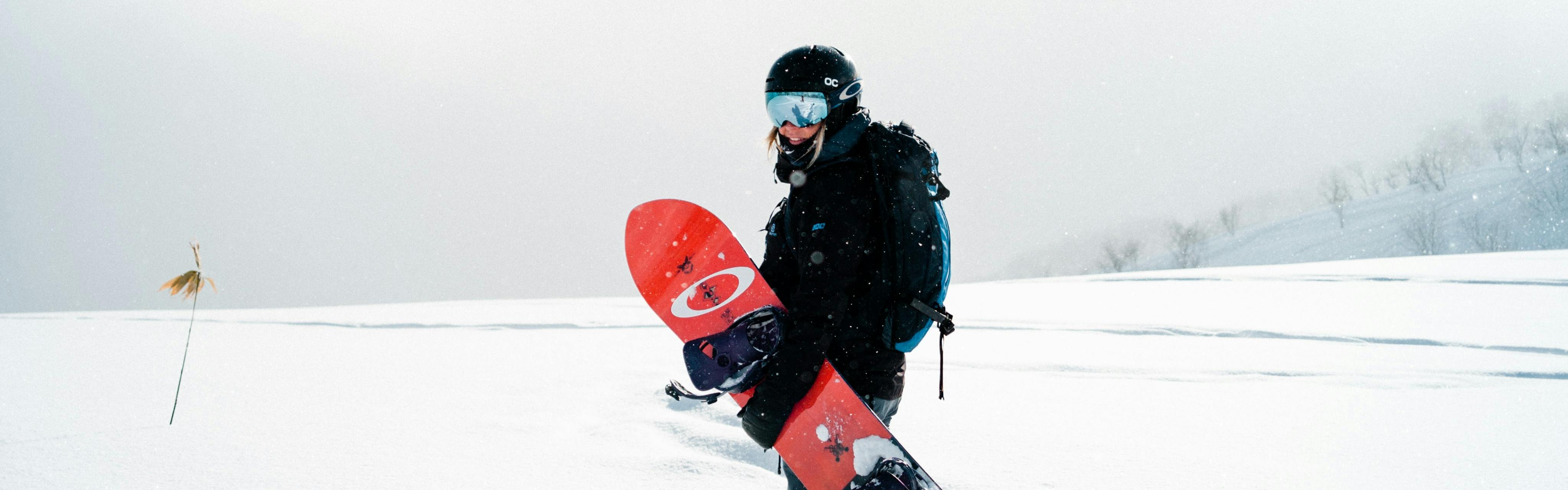 A woman stands and looks back at the camera while carrying a red snowboard and reflective goggles. The landscape around her is totally white and snowy.