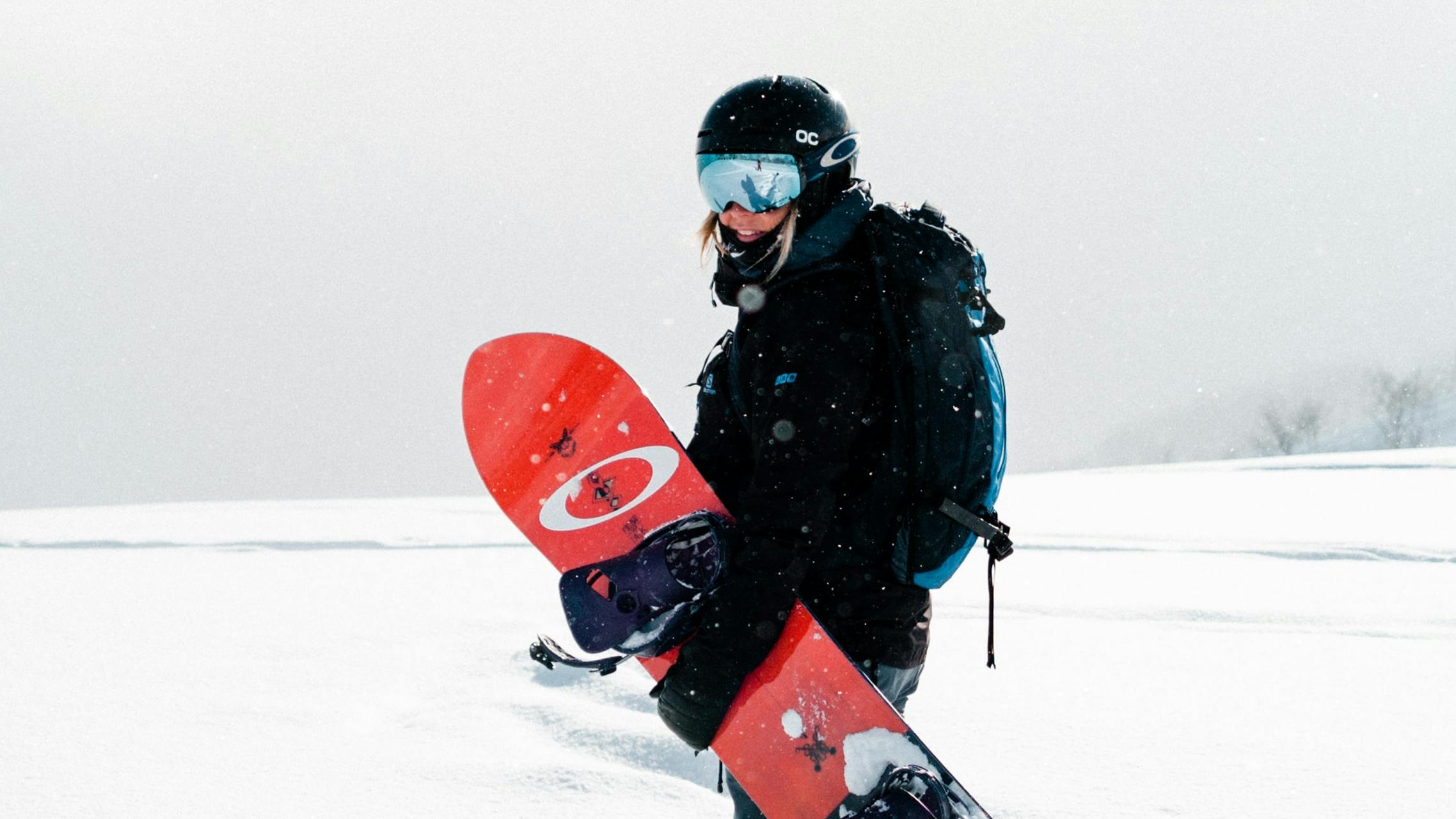 A woman stands and looks back at the camera while carrying a red snowboard and reflective goggles. The landscape around her is totally white and snowy.