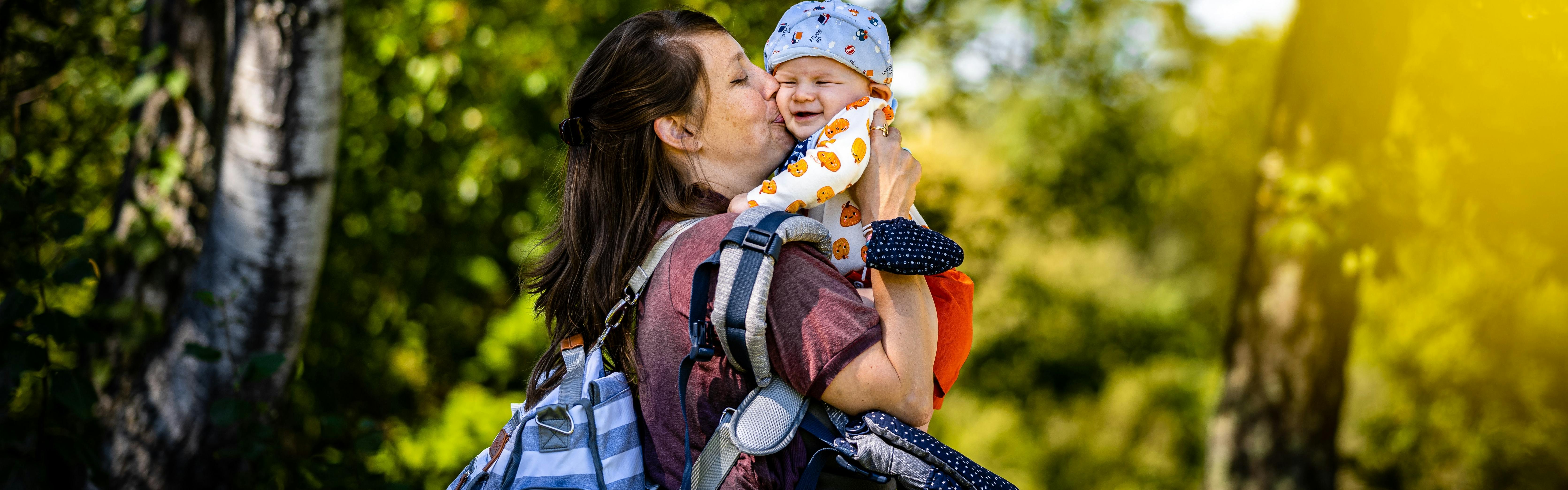 A woman kissing her baby. She has a backpack, diaper bag, and baby carrier on her shoulder.