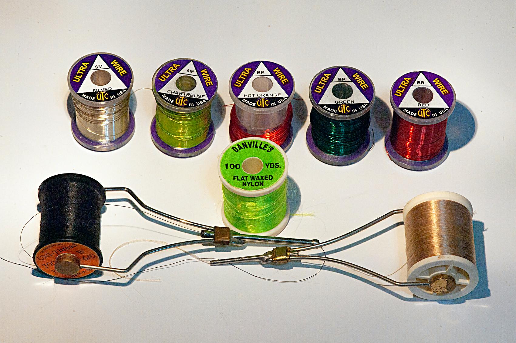 Six different types of thread for tying flies are pictured along with two wire bobbins holding two other kids of thread. The threads are a range of colors including green, red, tan, and black.