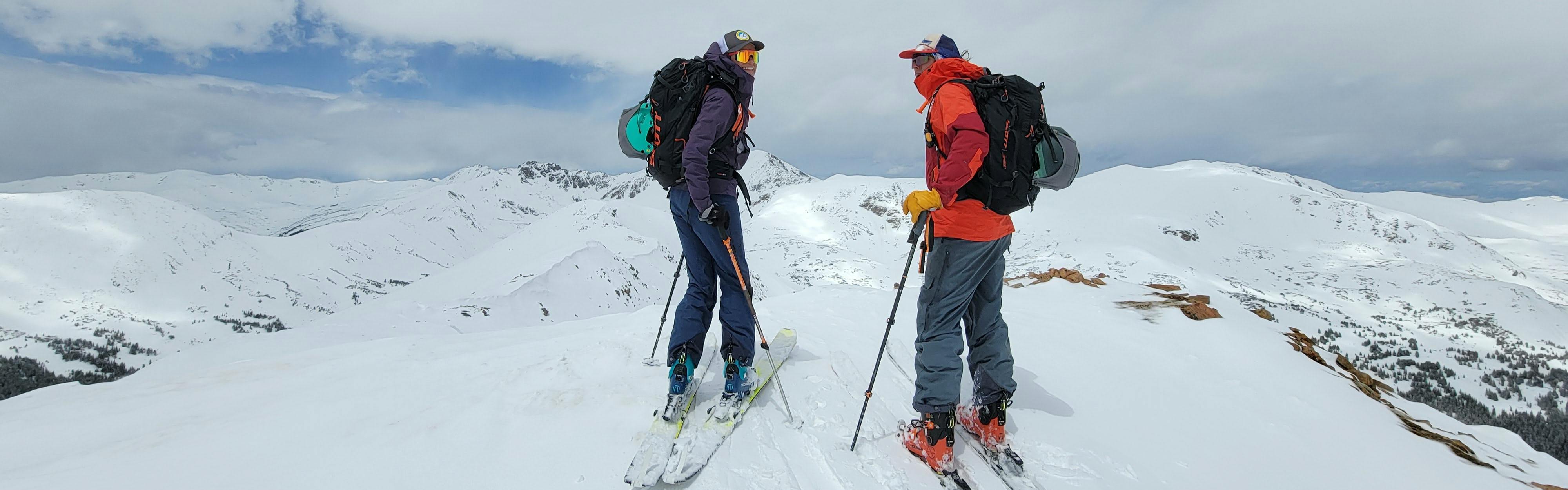 Two women are standing at the top of a snowy run with their backcountry ski gear.
