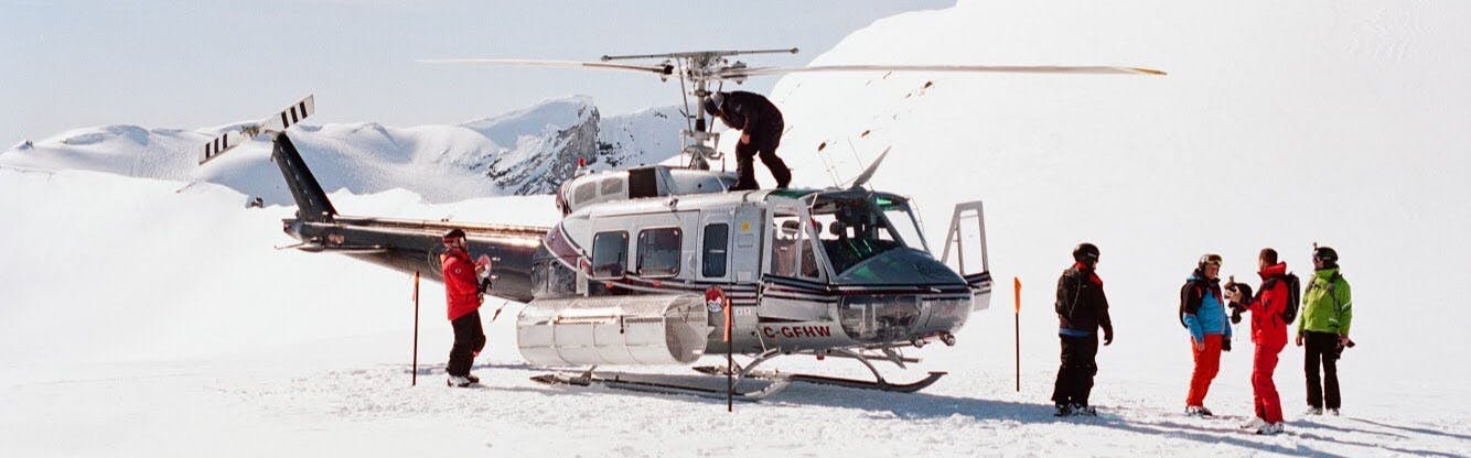 Several people with ski gear on stand in front of a helicopter that is parked in the snow. 