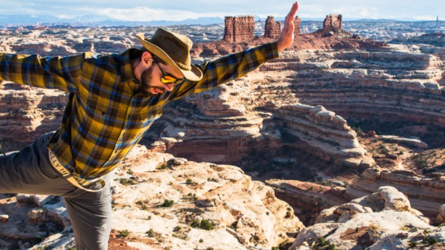 A man in a hat and a plaid shirt stretches his legs on top of a scenic outlook.