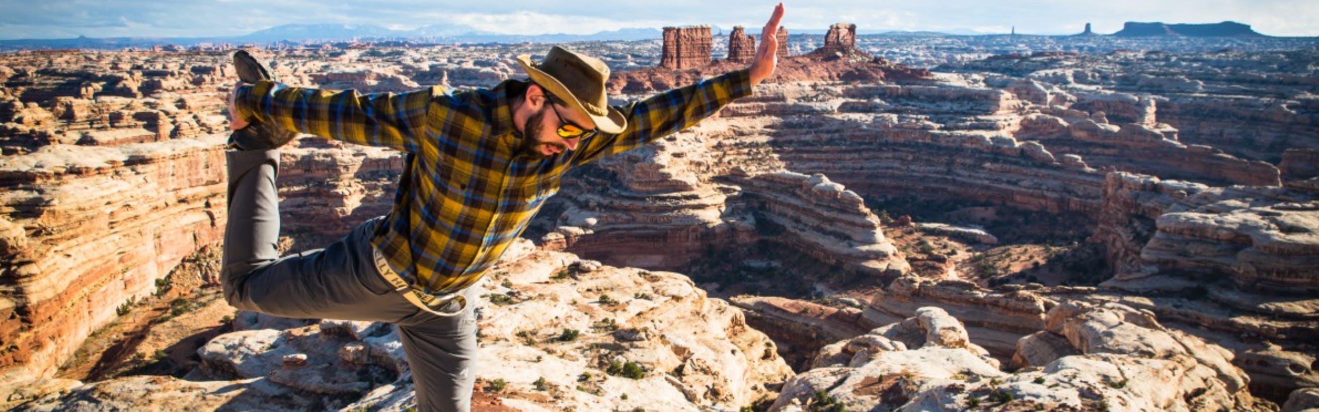 A man in a hat and a plaid shirt stretches his legs on top of a scenic outlook.