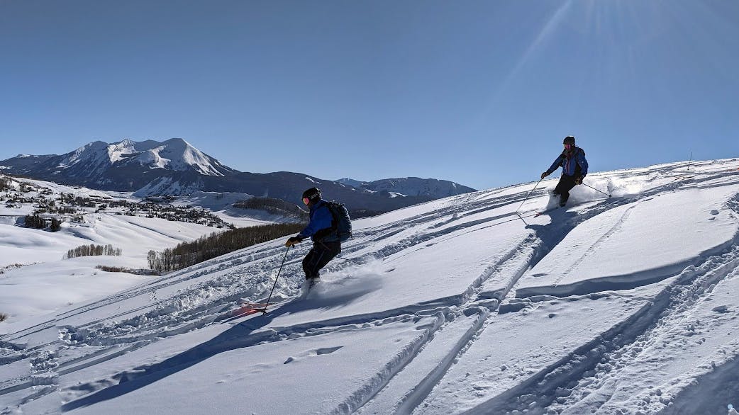 Four skiers skiing in a line down a snowy run. There are snowy mountains in the background.