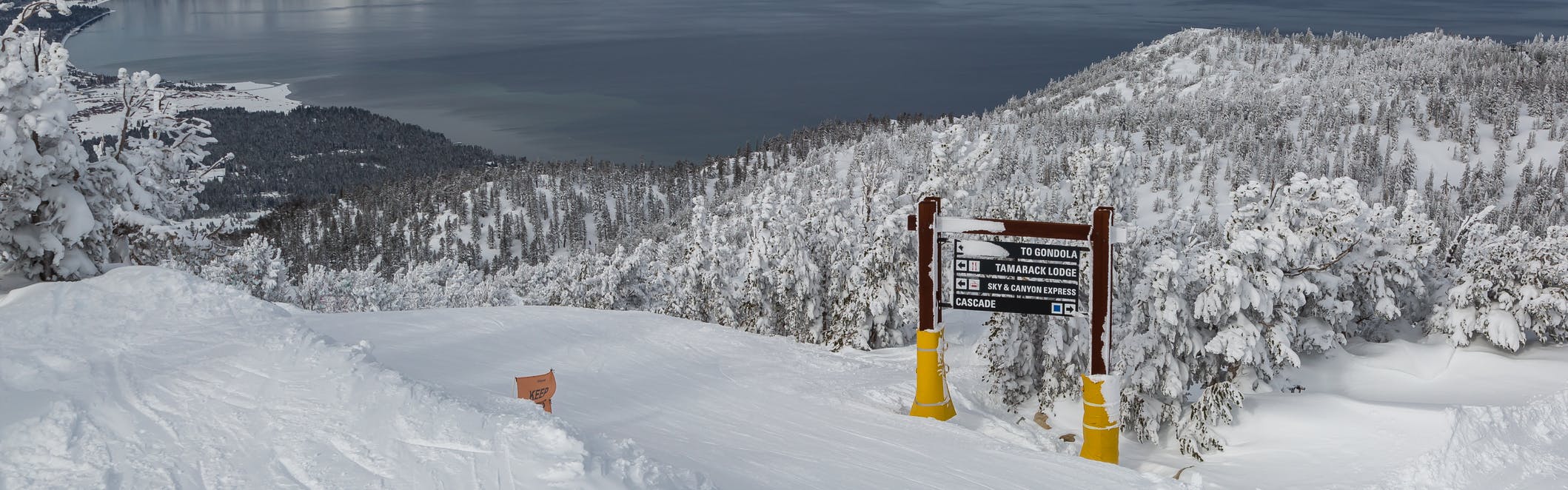 A sign listing ski runs sits on a snowy hill with trees and a lake in the background
