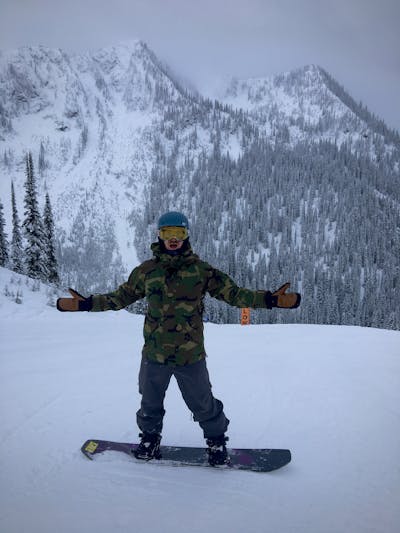 A man on the K2 Manifest Snowboard. He is standing on a run with his hands open. There is a mountain behind him. 