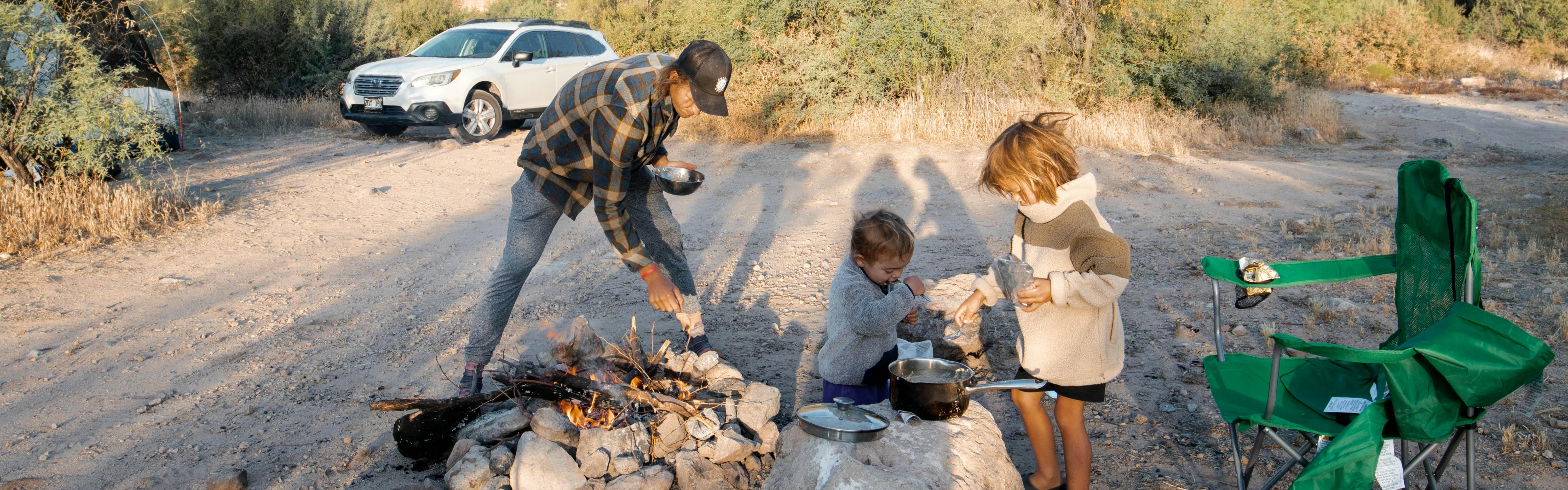 An adult stokes a campfire as two kids put food in a pot. There is a camp chair sitting next to them. 