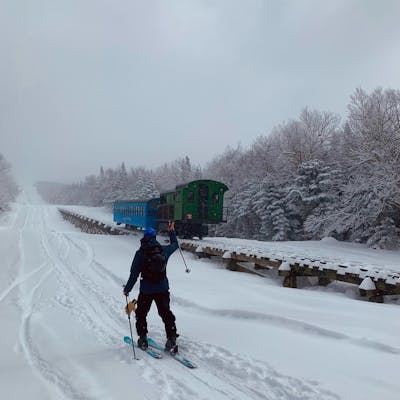 A skier walking on his skis waves at a train. There is snow all around. 