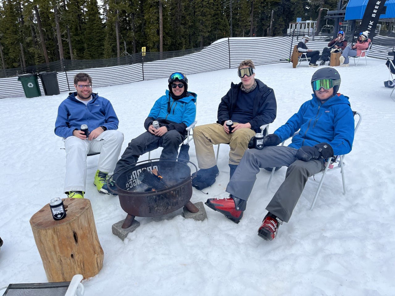 Four skiers in ski gear are sitting in chairs around a fire. There is snow all around them and a ski resort in the background. 