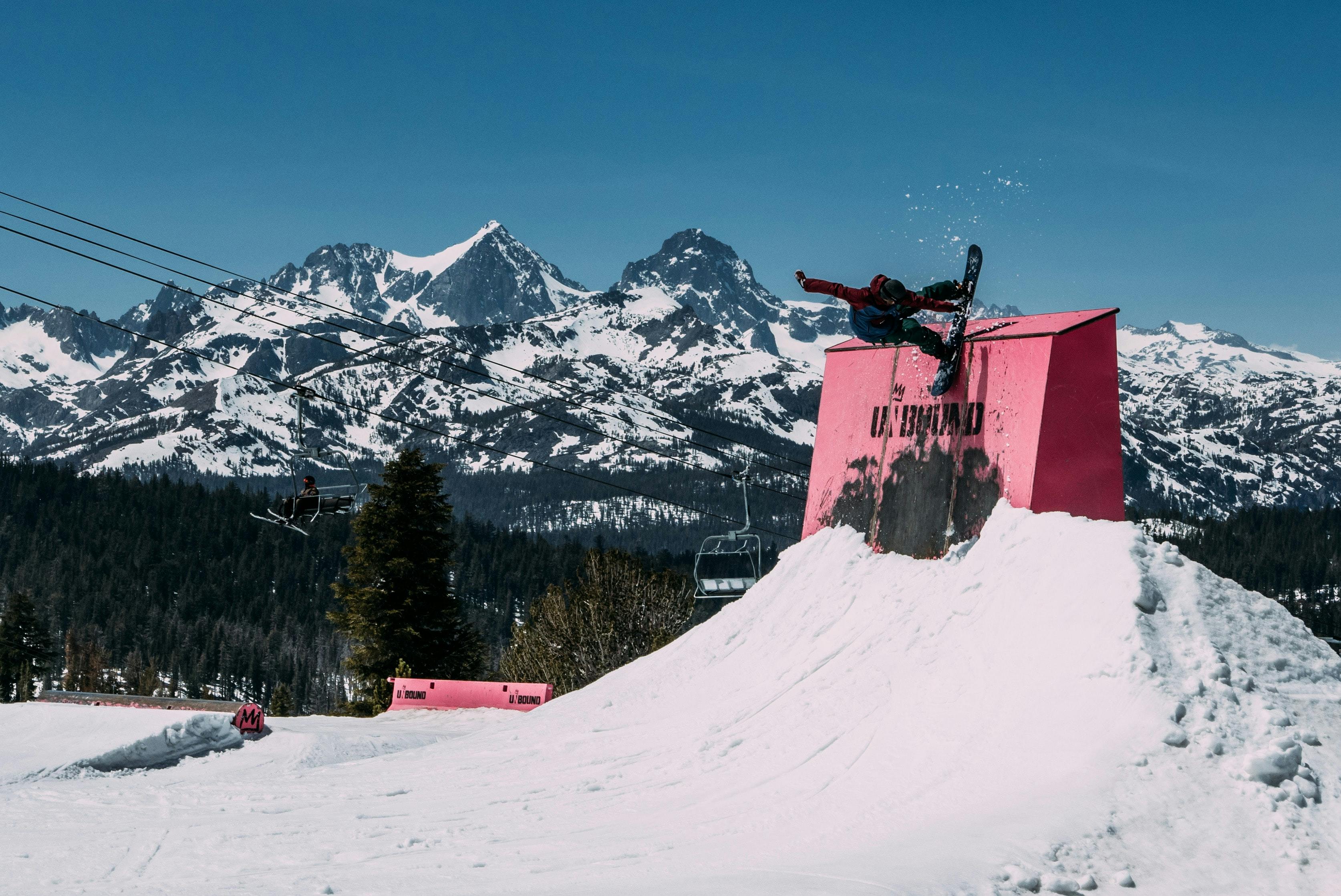 A snowboarder executes a trick on a red half wall with mountains in the background