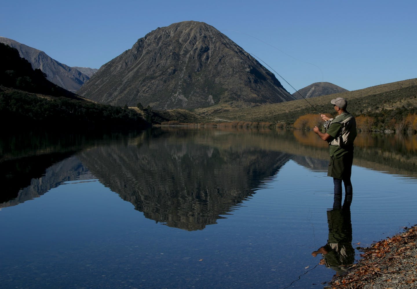 A man fly fishes on a lake in NZ that is still and perfectly reflects the surrounding hills.