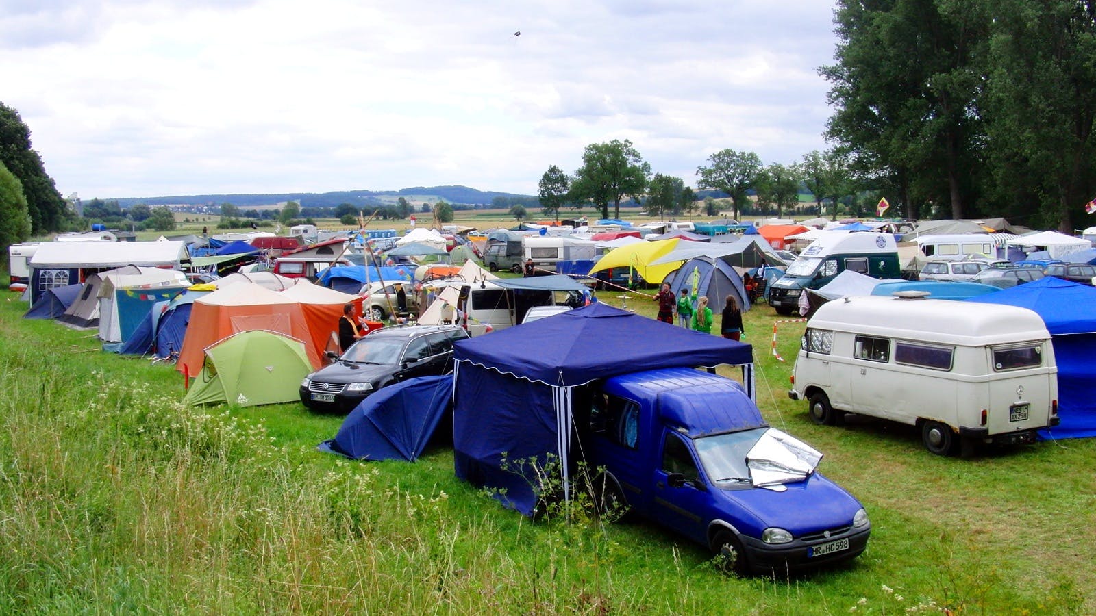 Cars and tents share the space in a festival camping ground. Pop-up canopies are used to create shade.