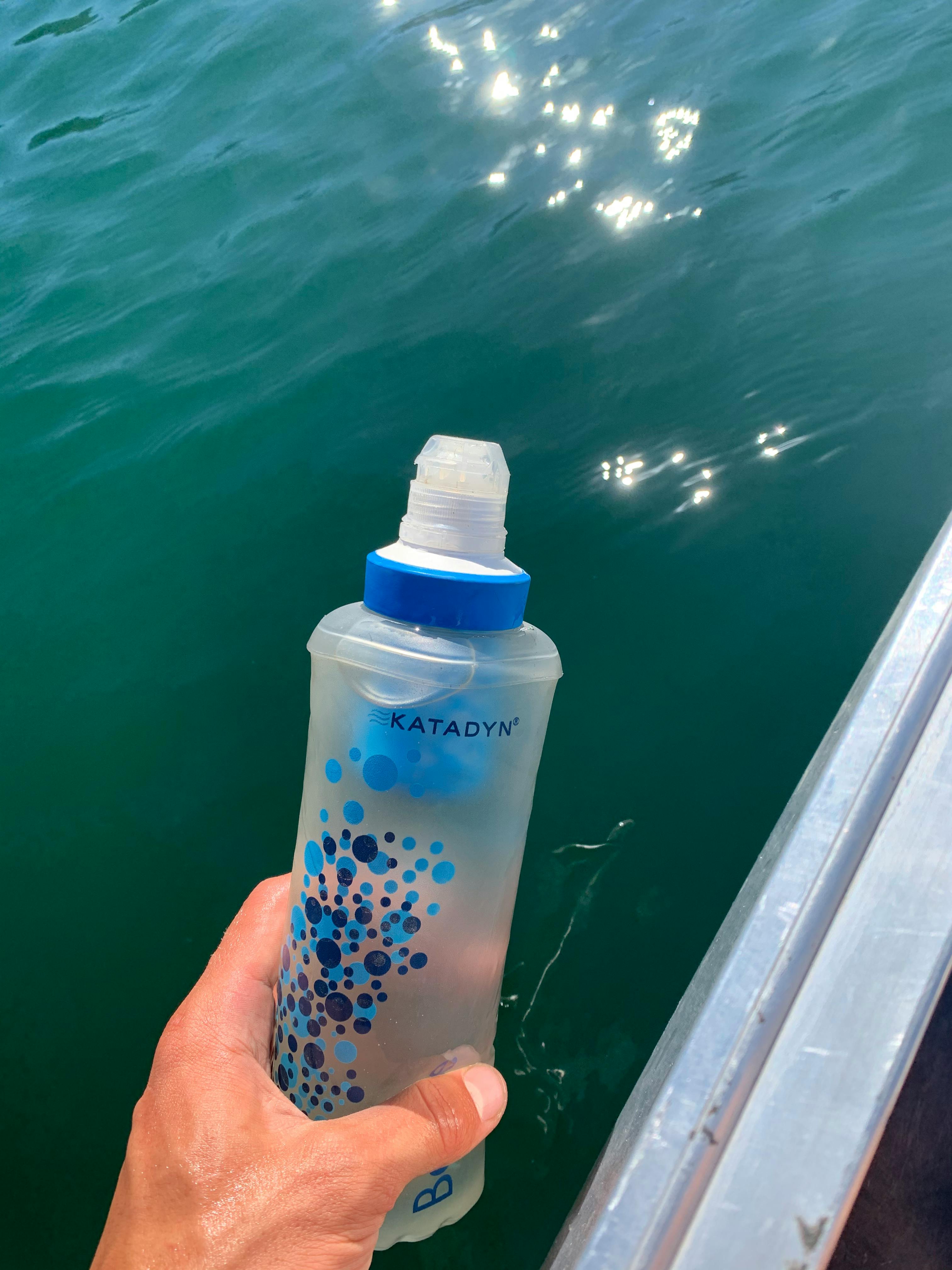A person's hand holds a water bottle filter over some blue-green water
