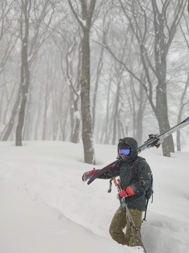 A man is holding a pair of skis over his shoulder. It is snowing and he is walking up a trail.