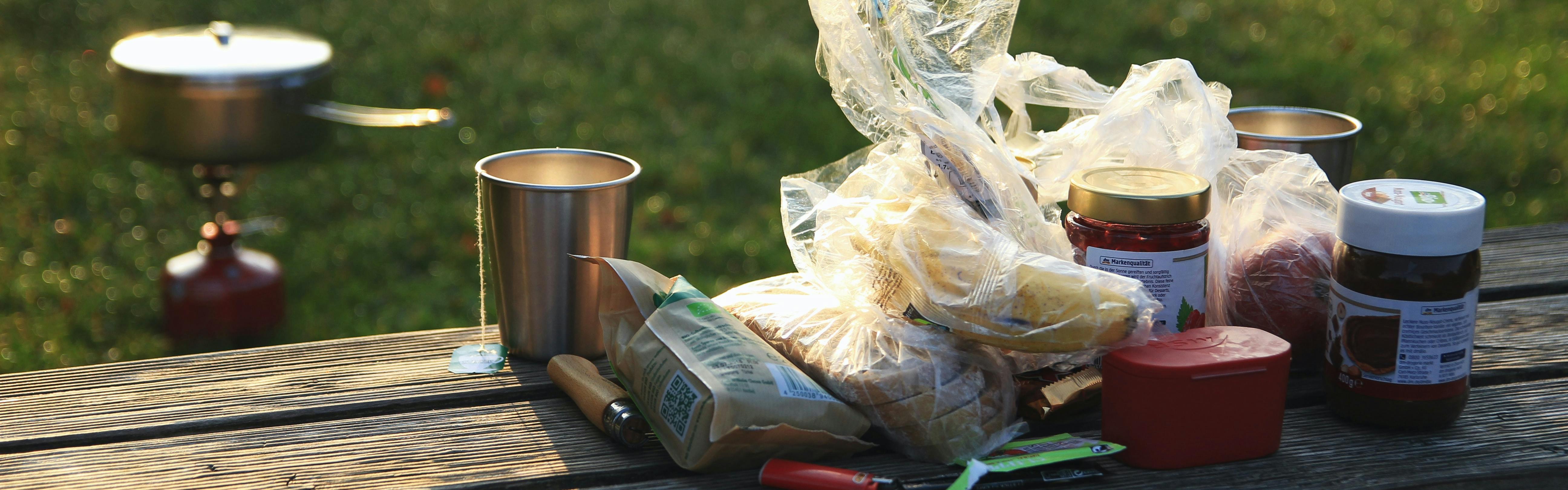 A bunch of breakfast ingredients sit on a wooden picnic table. In the background, someone heats water on a backpacking stove. 