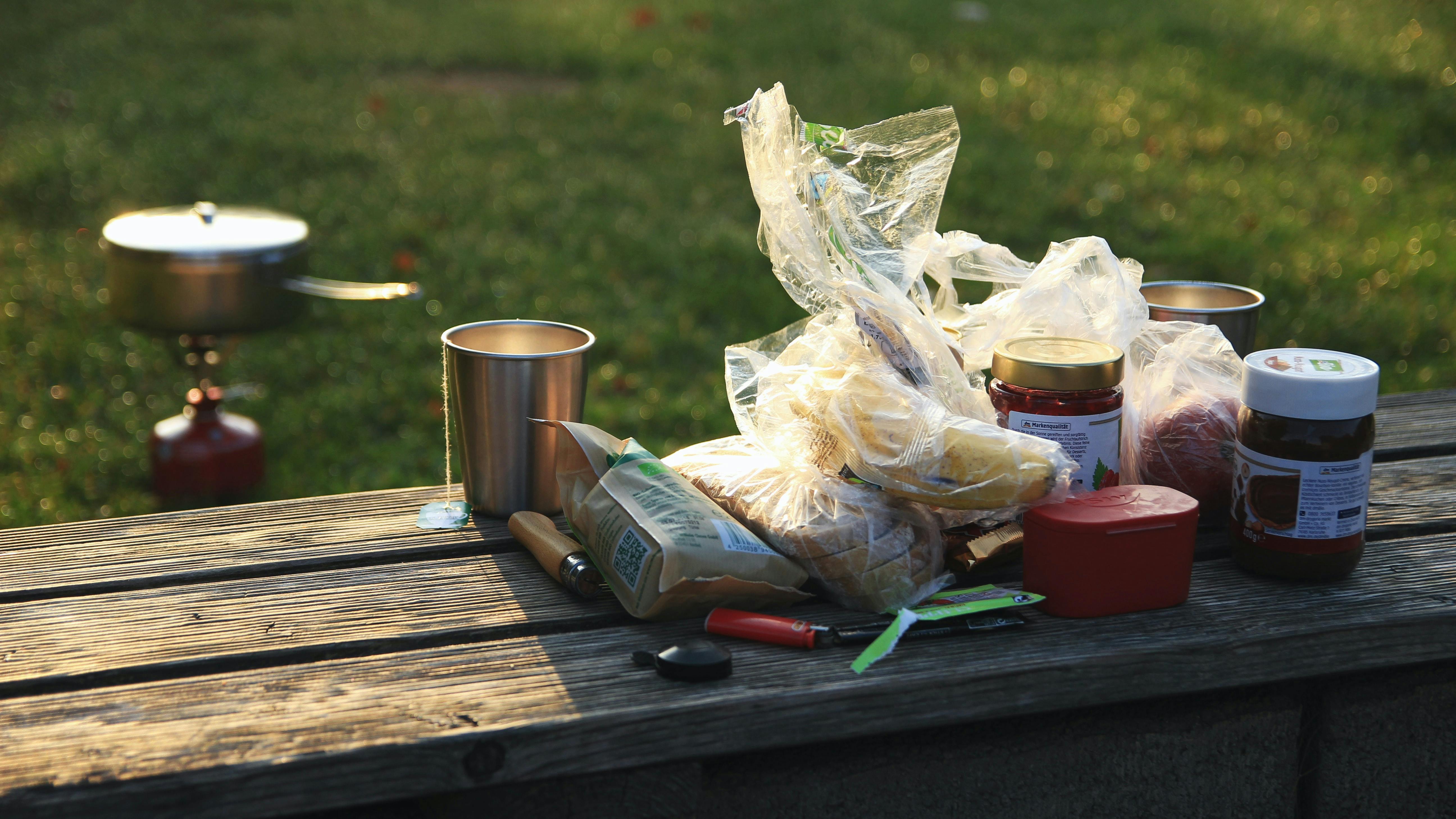 A bunch of breakfast ingredients sit on a wooden picnic table. In the background, someone heats water on a backpacking stove. 