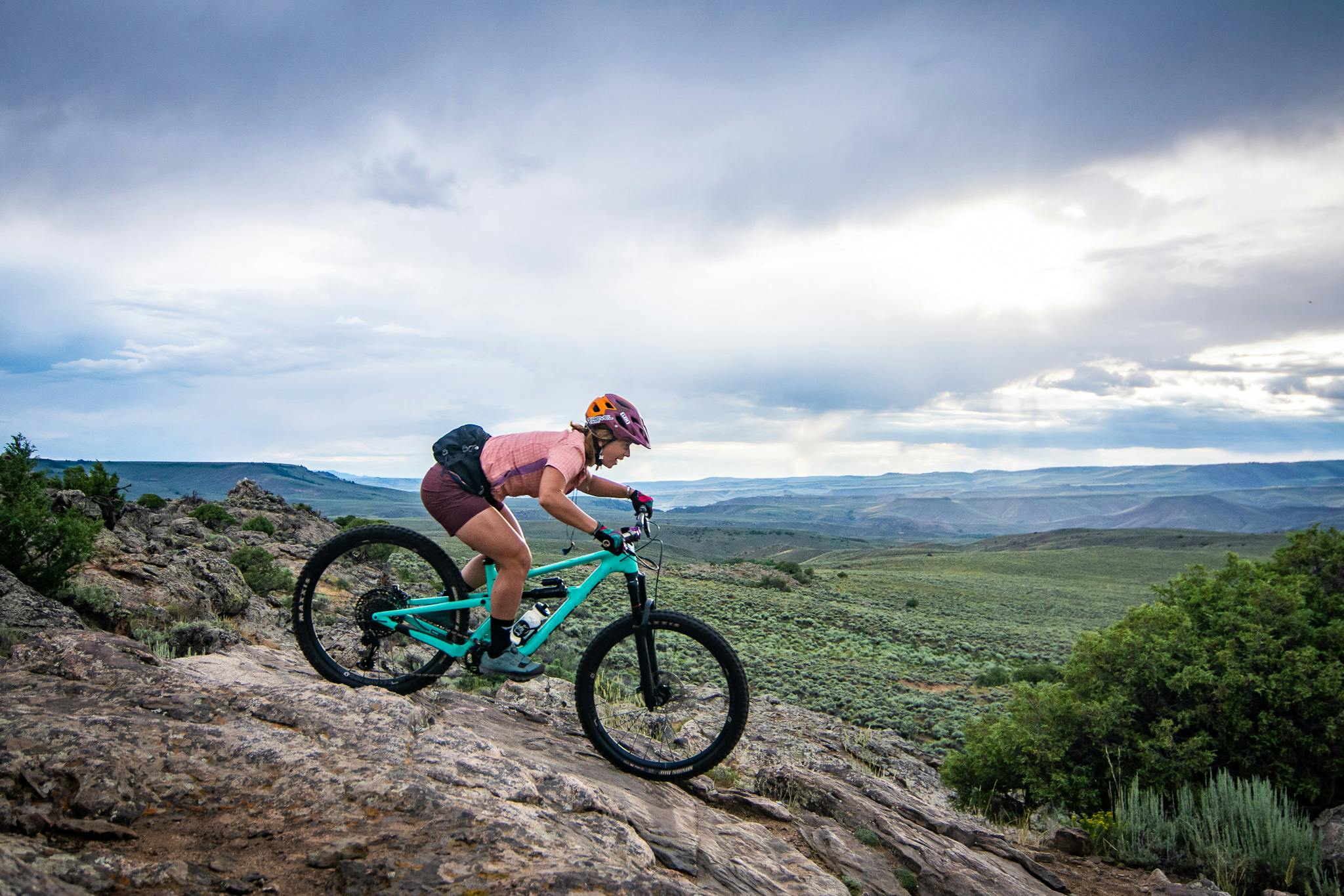 A woman mountain biking down a rocky slope with an expansive view of hills in the distance.