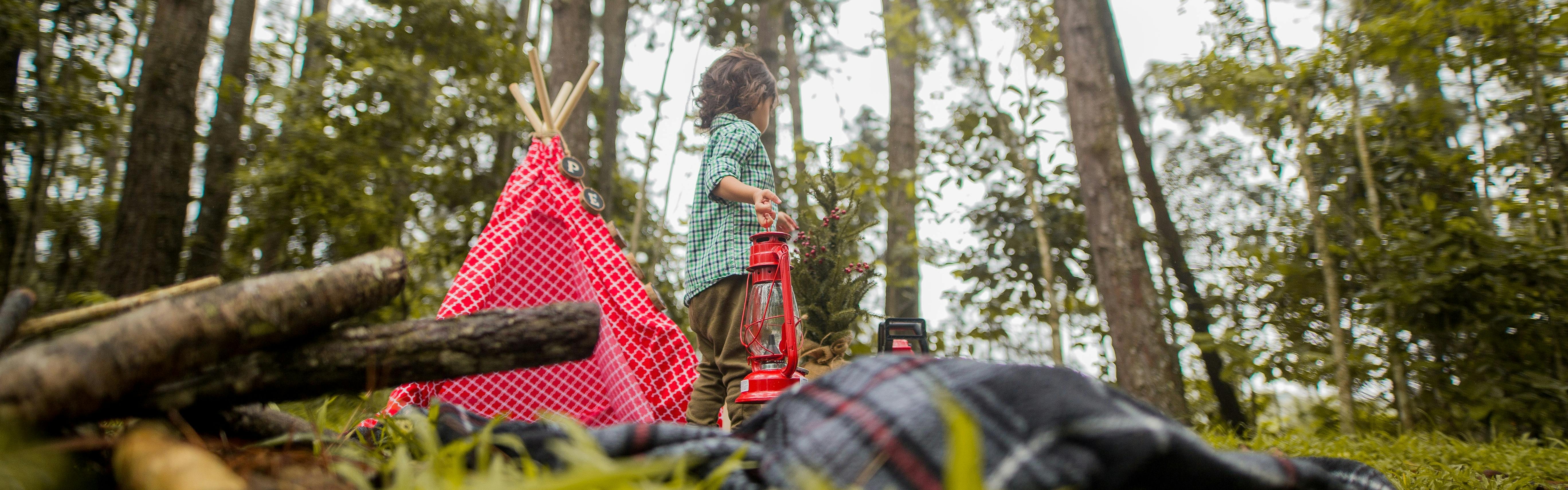 A toddler in a plaid shirt carries a red lantern and stands next to a small red teepee-style tent in a forest