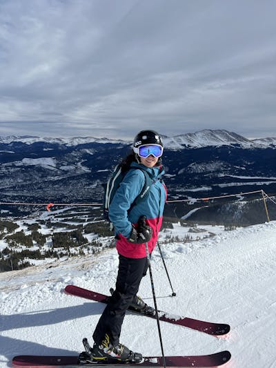 A woman standing on skis at the top of a ski run. There are mountains in the distance.