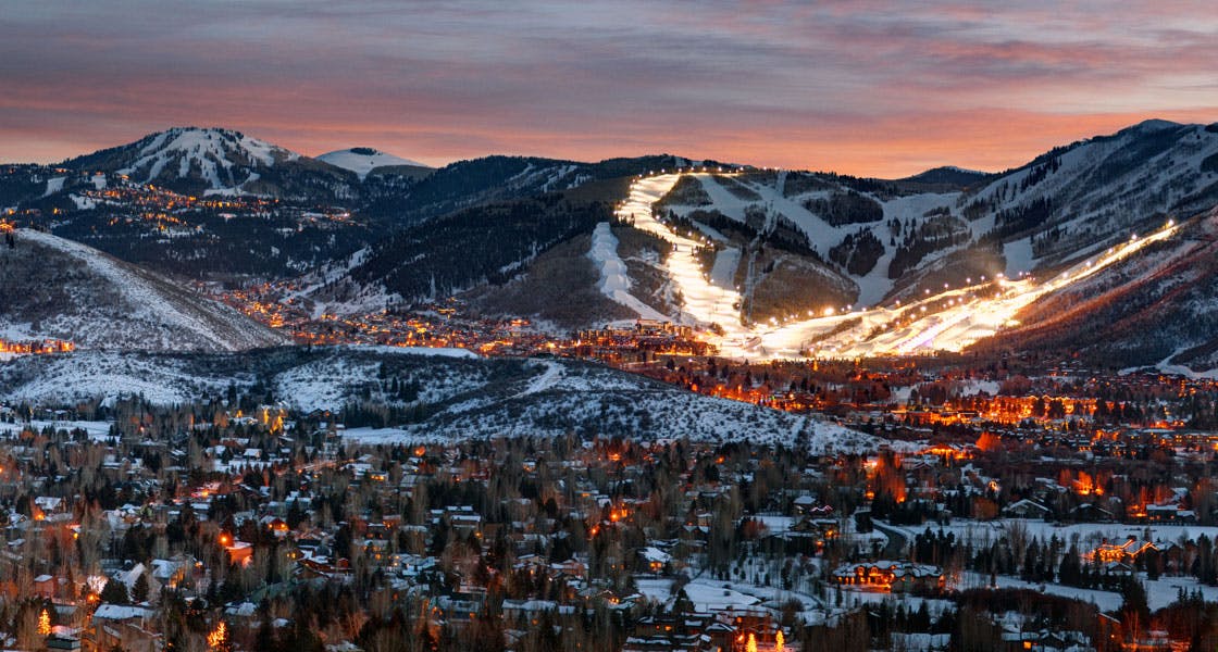 A landscape of Park City taken at sunset with the ground snowy and the city lights lit.