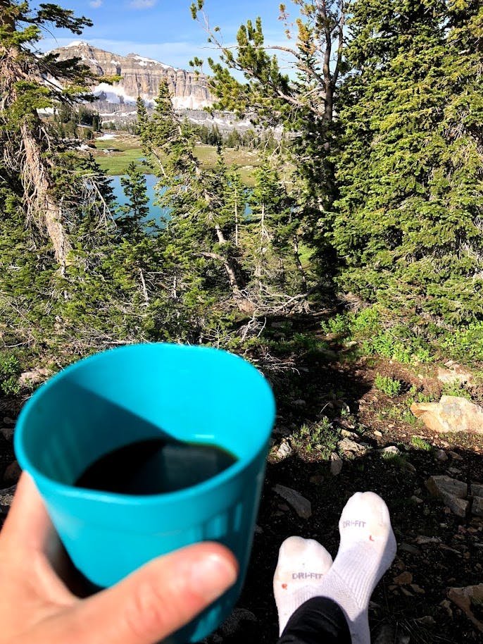 The author takes a picture of her hand holding a blue cup full of coffee with a view of pine trees, a lake, and a peak in the distance. 